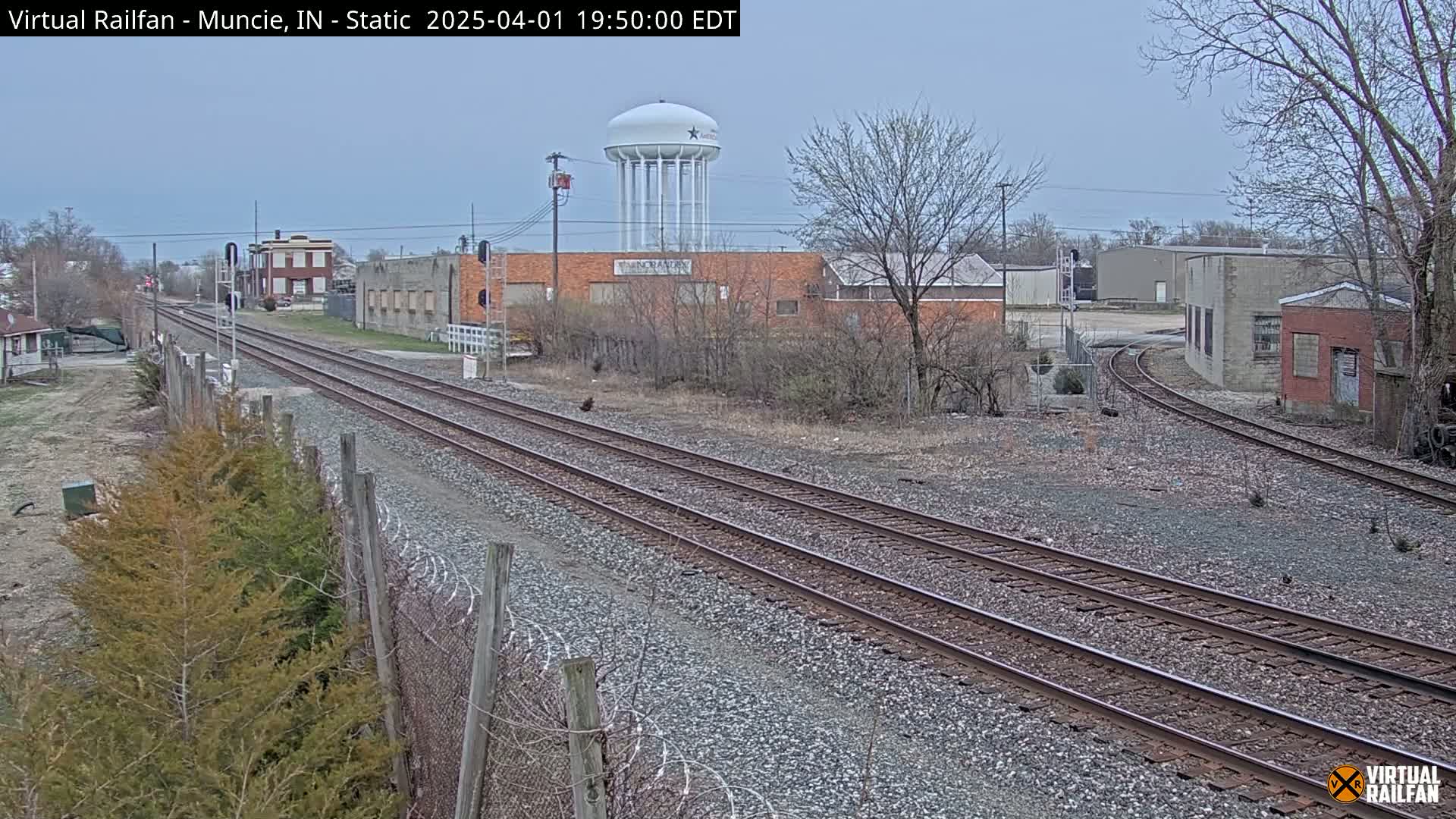 A view of railroad tracks stretching into the distance, passing industrial buildings and a water tower under an overcast sky.
