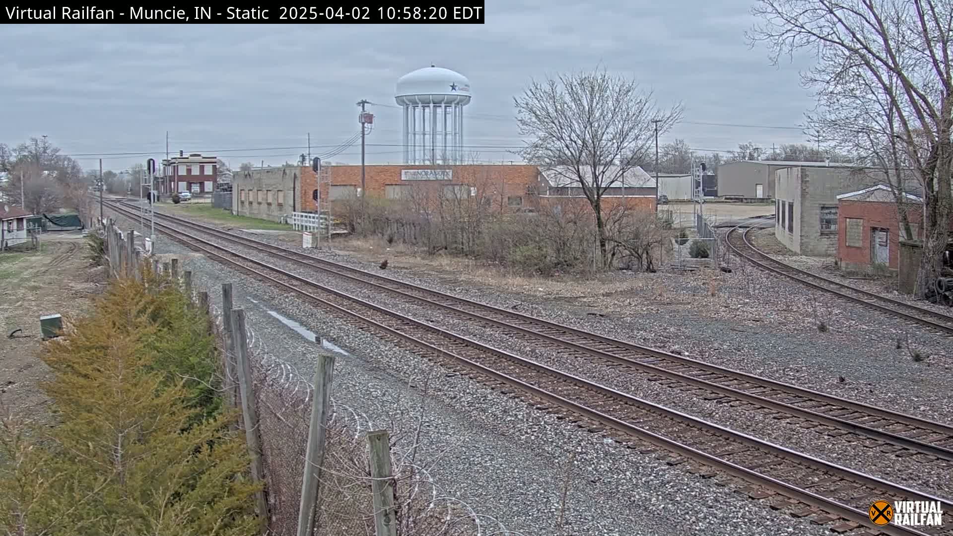 A view of railroad tracks extending into the distance under an overcast sky, passing industrial buildings and a water tower.