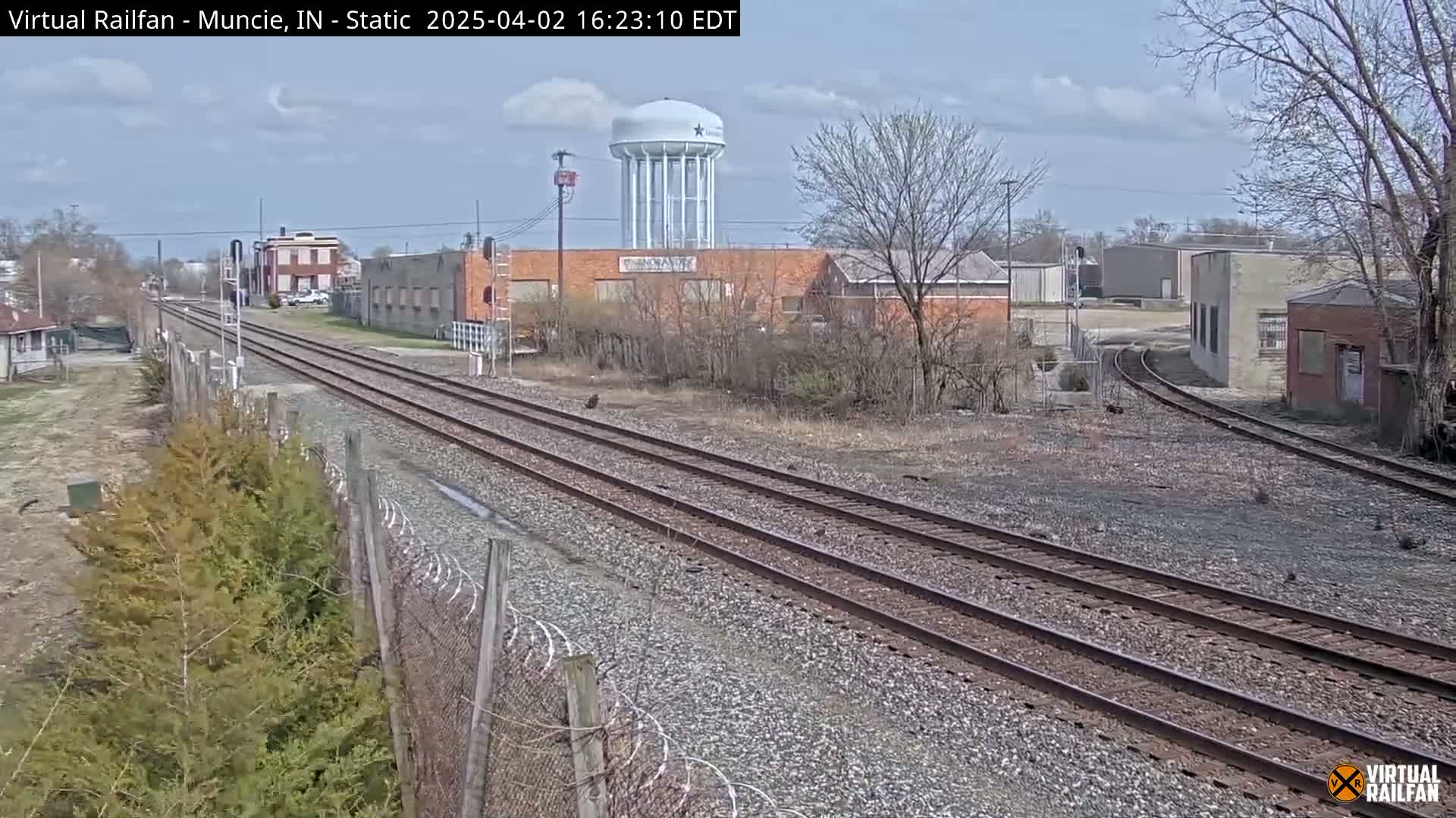Railroad tracks extend into the distance past industrial buildings and a water tower under a partly cloudy sky.
