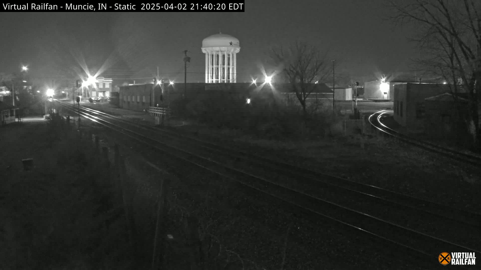 A nighttime view of railroad tracks leading toward a water tower and industrial buildings under a dark sky.