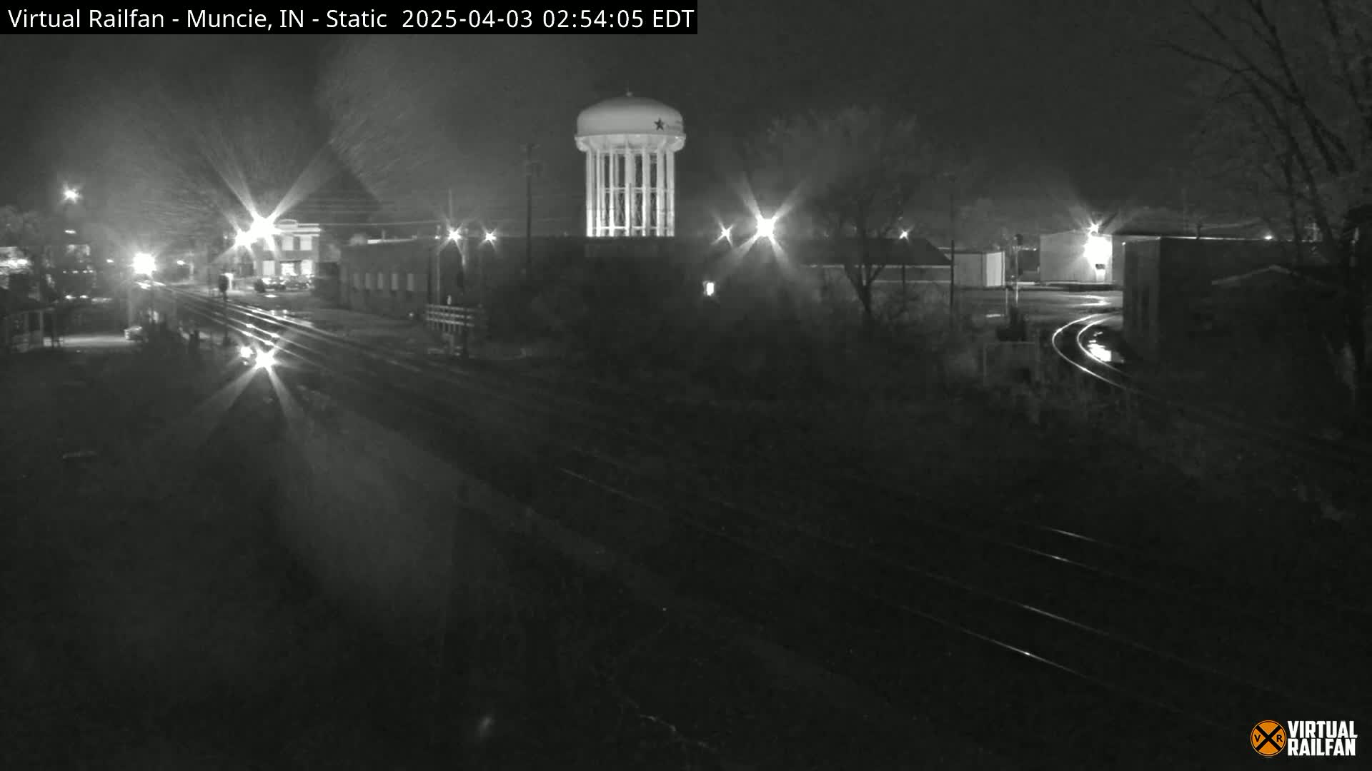 A nighttime, low-light view of a water tower and railroad tracks, with several buildings visible in the background.