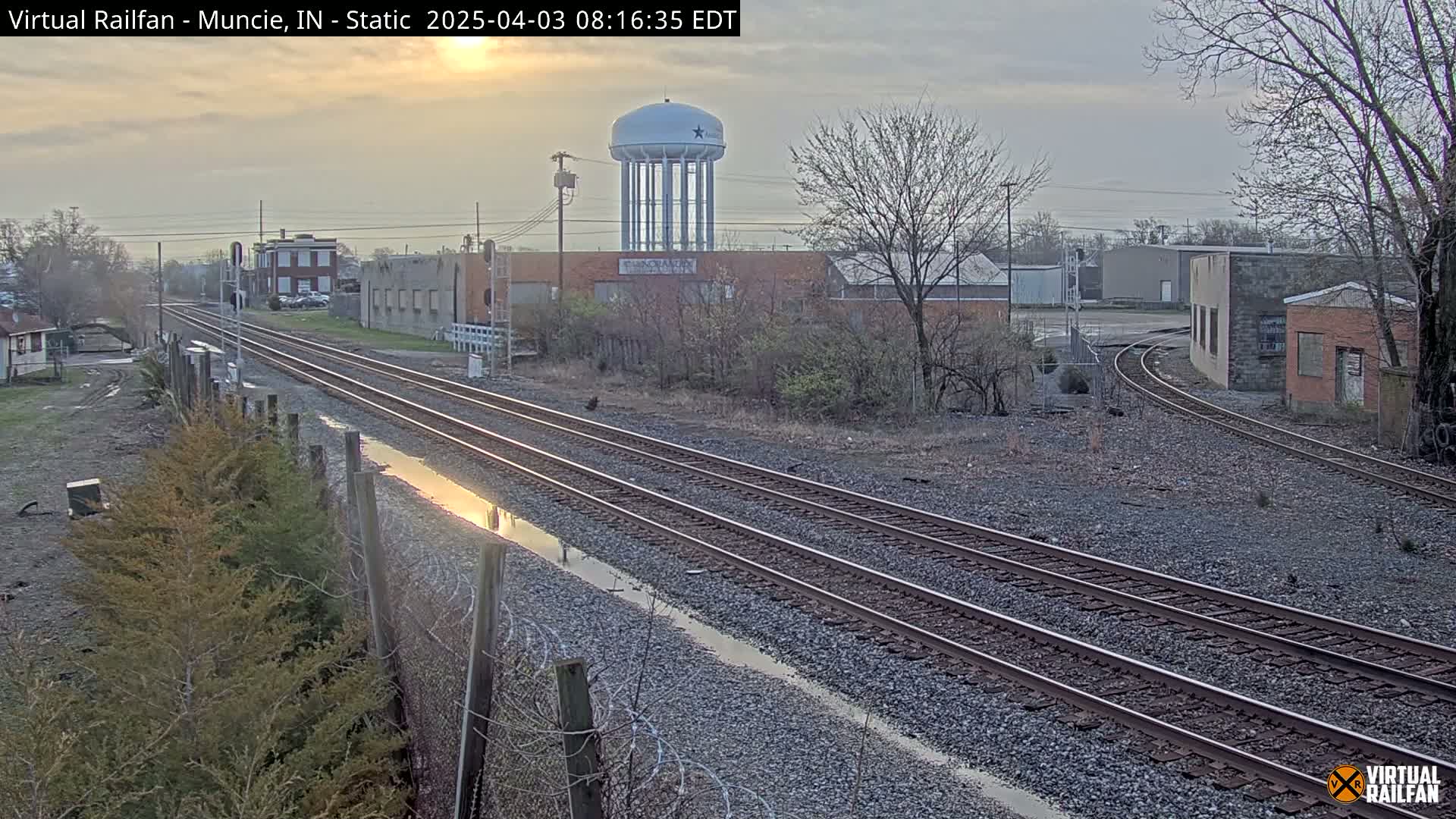 Three sets of train tracks extend into the distance under an overcast sky, past industrial buildings and a water tower.