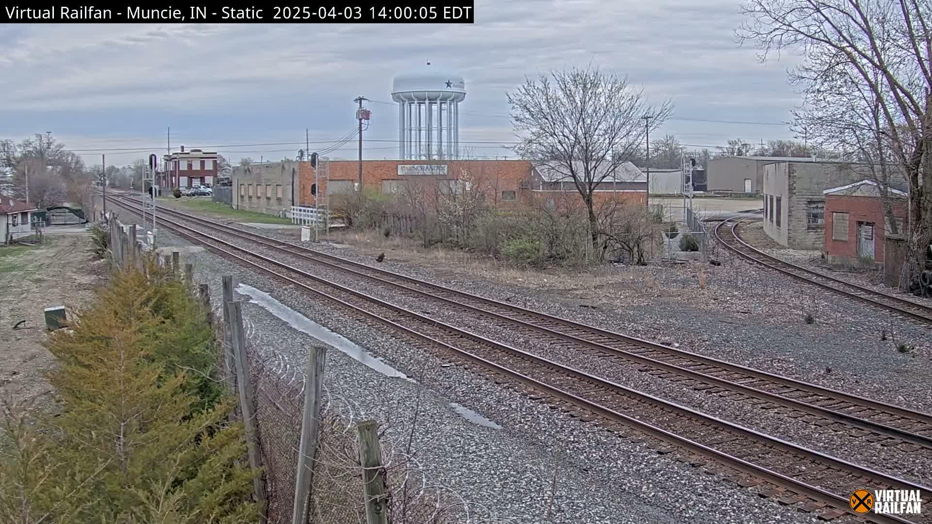 A view of railroad tracks on an overcast day, curving past industrial buildings and a water tower, with bare trees and shrubs in the foreground.