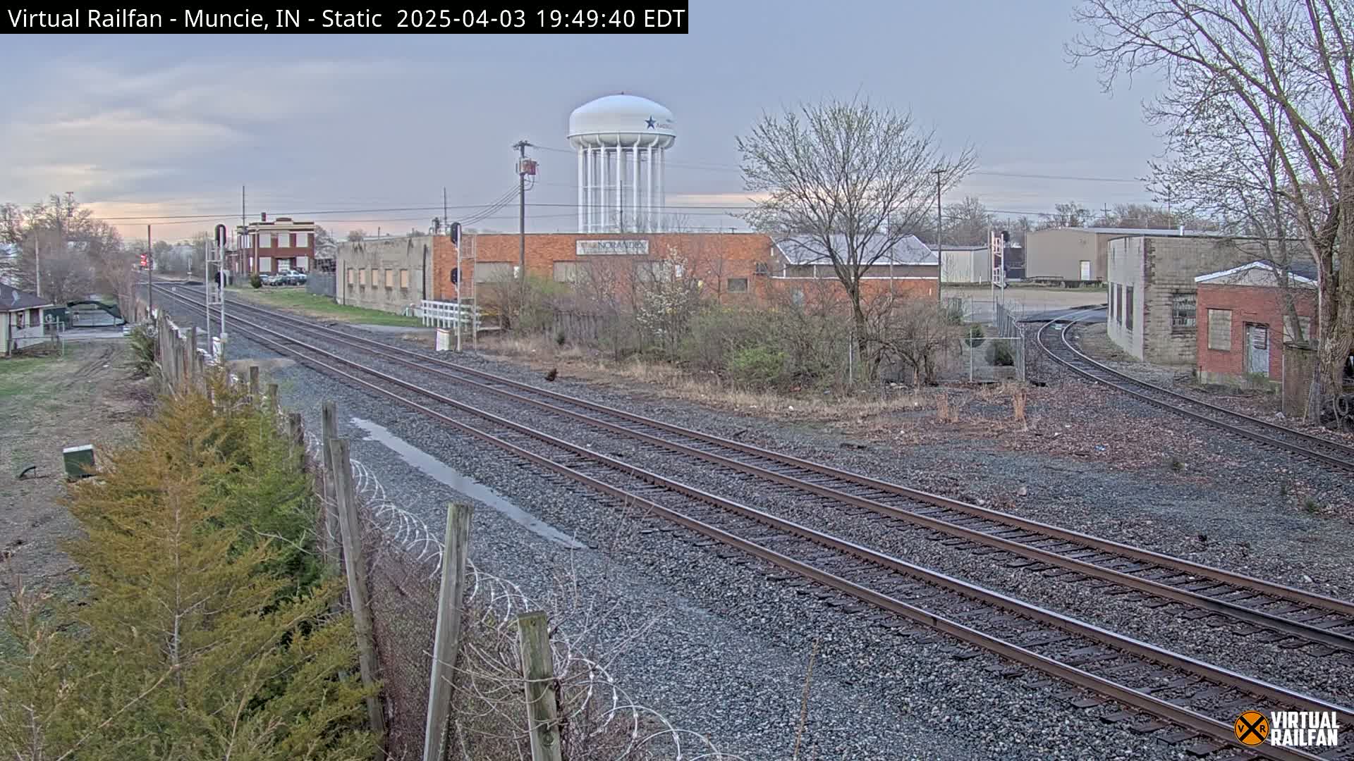 Railroad tracks extend into the distance past industrial buildings and a water tower under a cloudy sky.