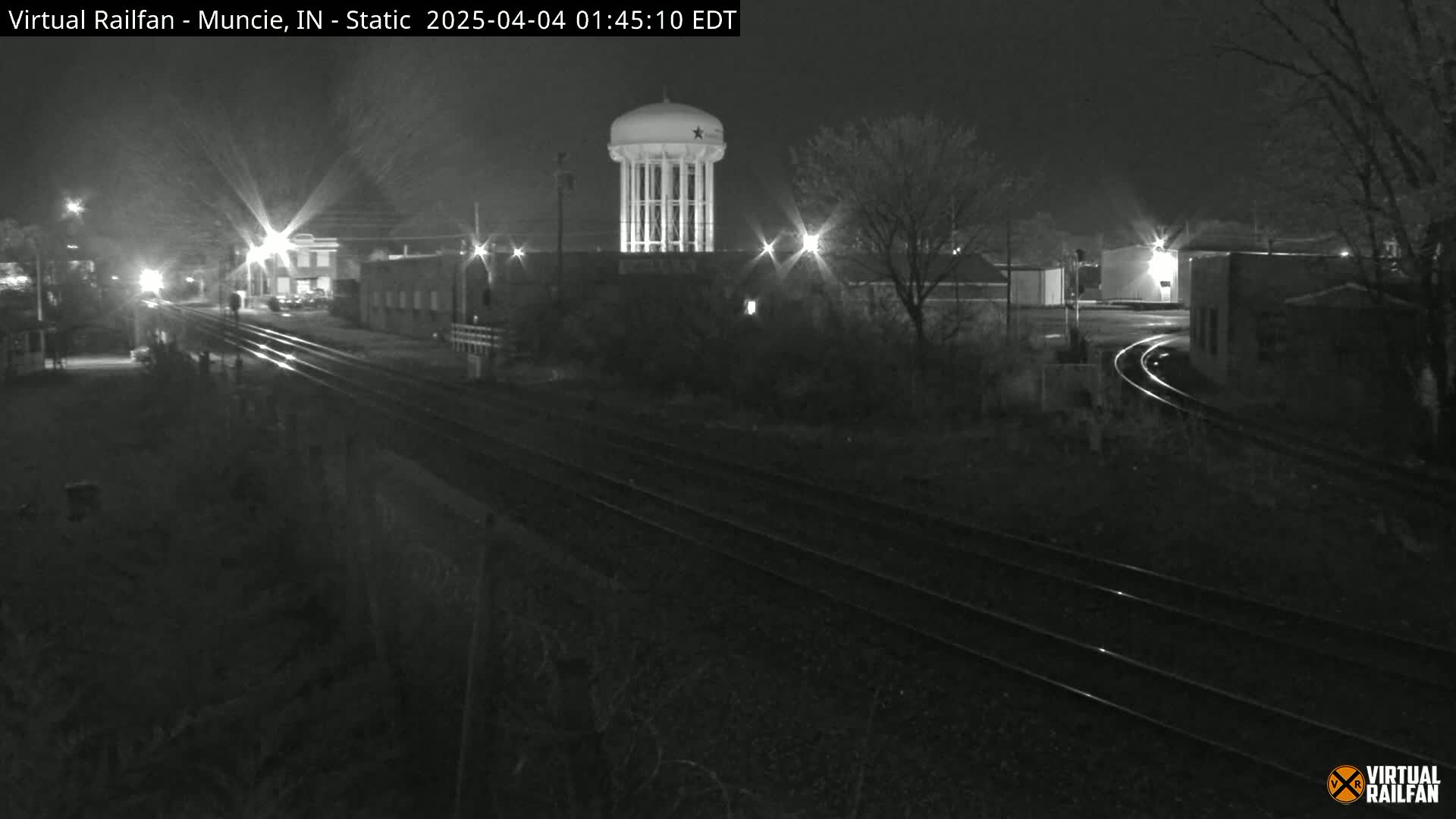 A nighttime view of train tracks and a water tower, illuminated by streetlights, under a dark sky.