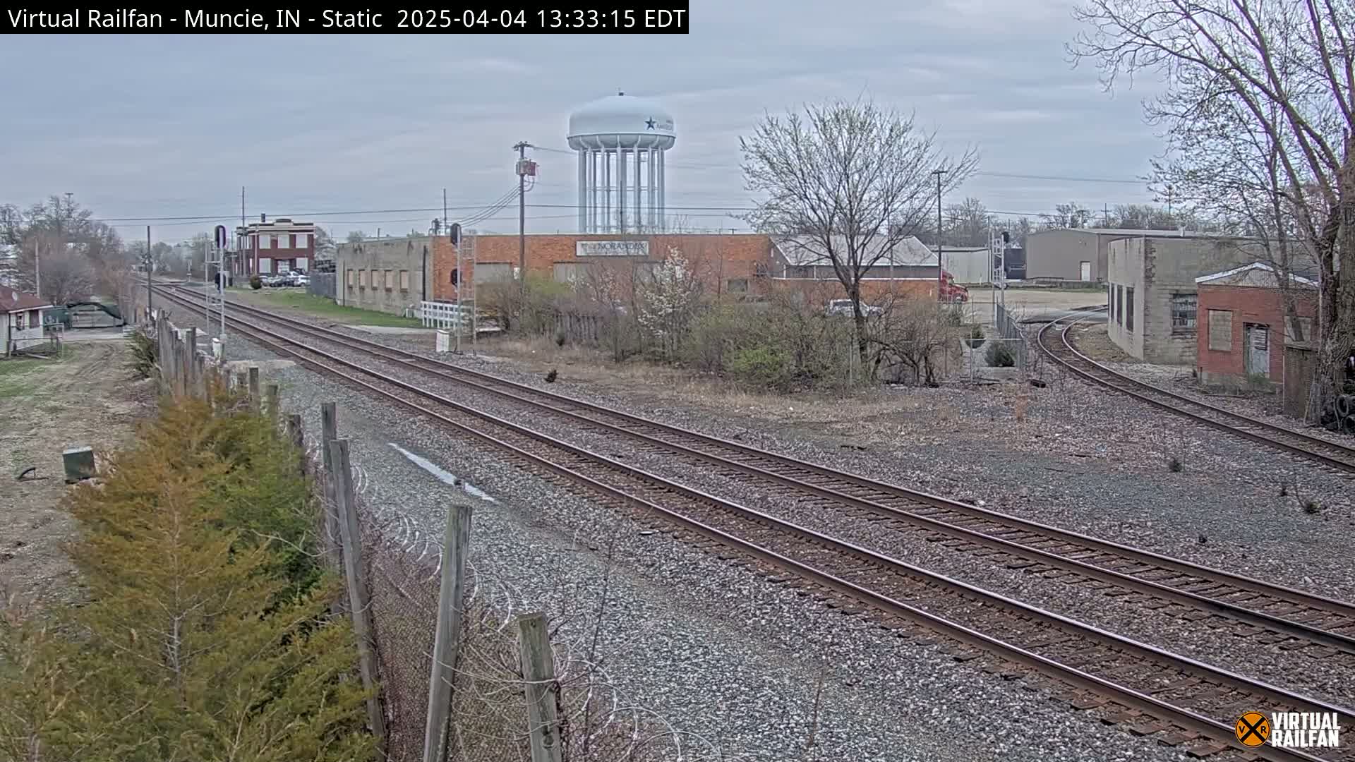 A view of railroad tracks extending into the distance, passing industrial buildings and a water tower under an overcast sky.