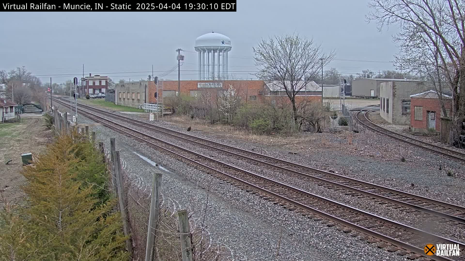 A view of railroad tracks extending into the distance on an overcast day, passing industrial buildings and a water tower.