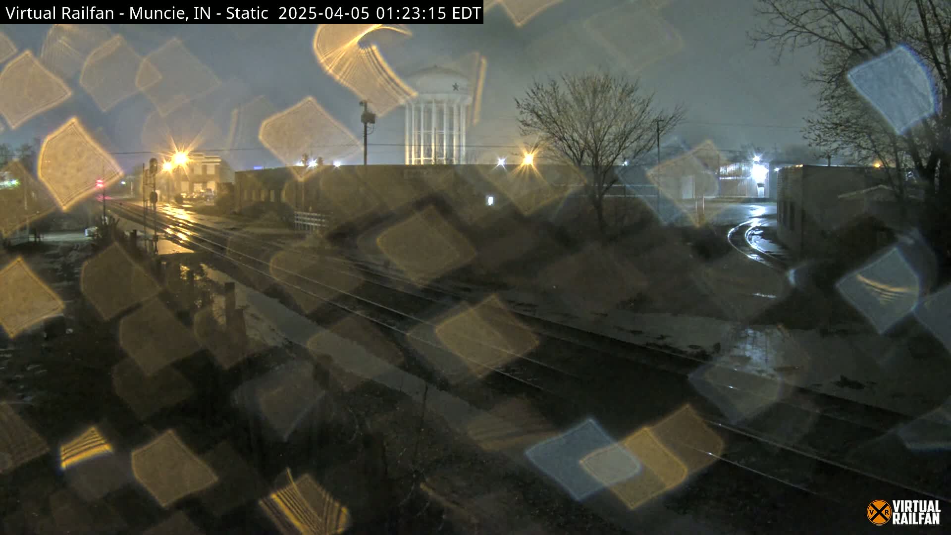 A nighttime view of rain-slicked railroad tracks and city buildings, seen through a rain-covered window.