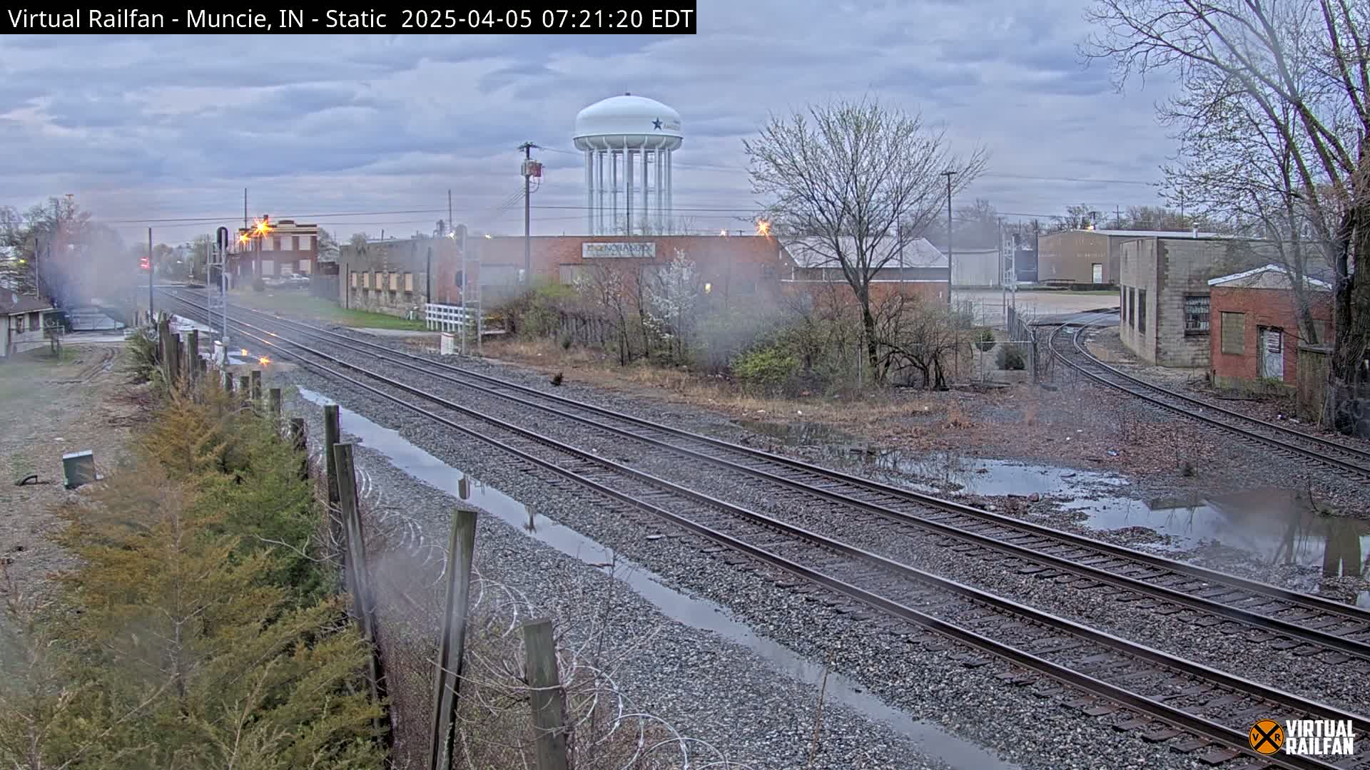 A view of railroad tracks on an overcast day with puddles of water along the tracks and in the surrounding area, industrial buildings in the background, and a water tower visible in the distance.