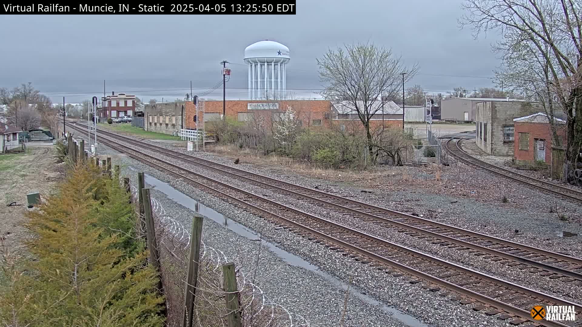 Overcast skies and wet ground surround railway tracks that curve around industrial buildings and a water tower.