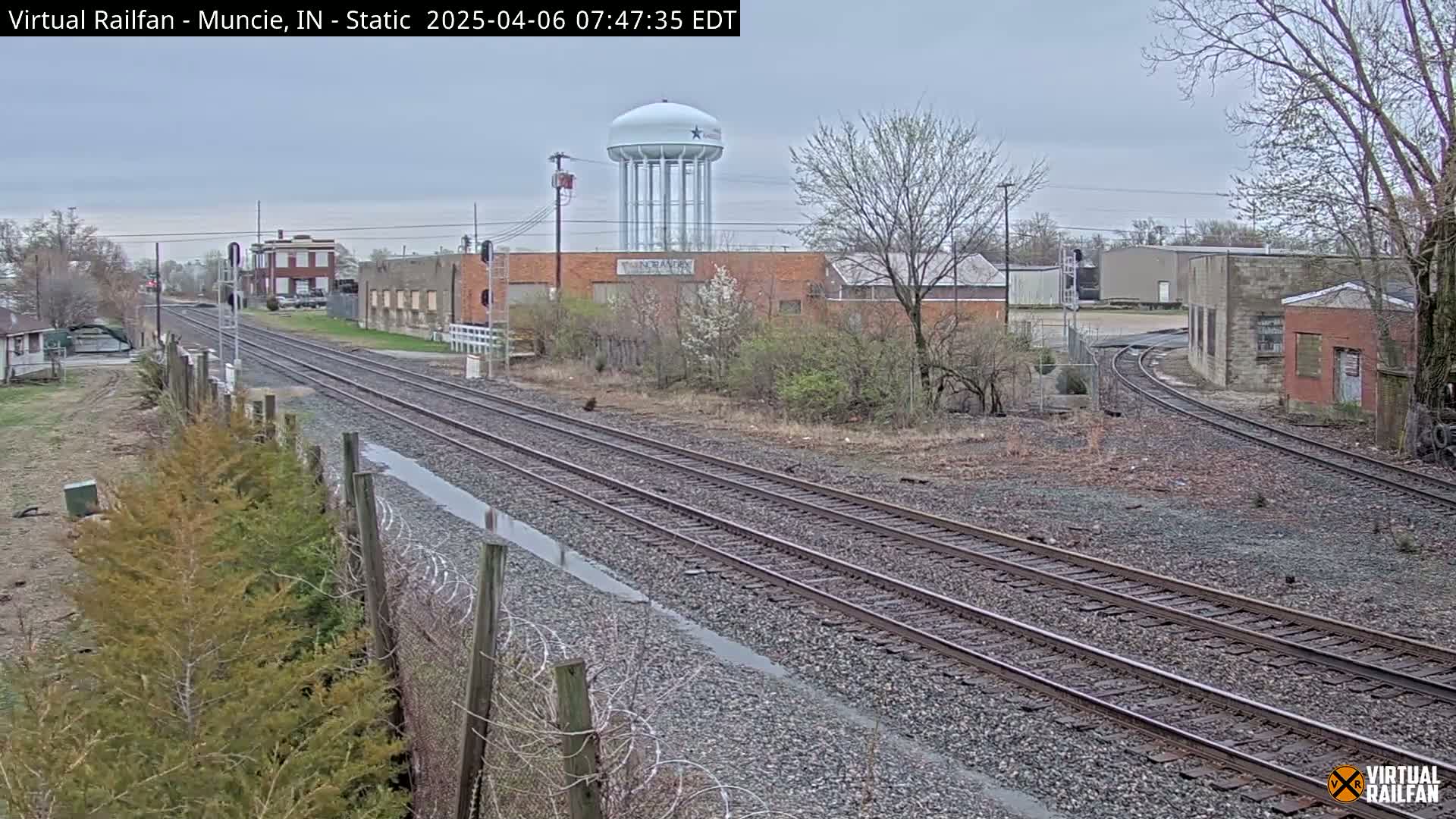 Multiple sets of train tracks extend into the distance under an overcast sky, passing industrial buildings and a water tower.