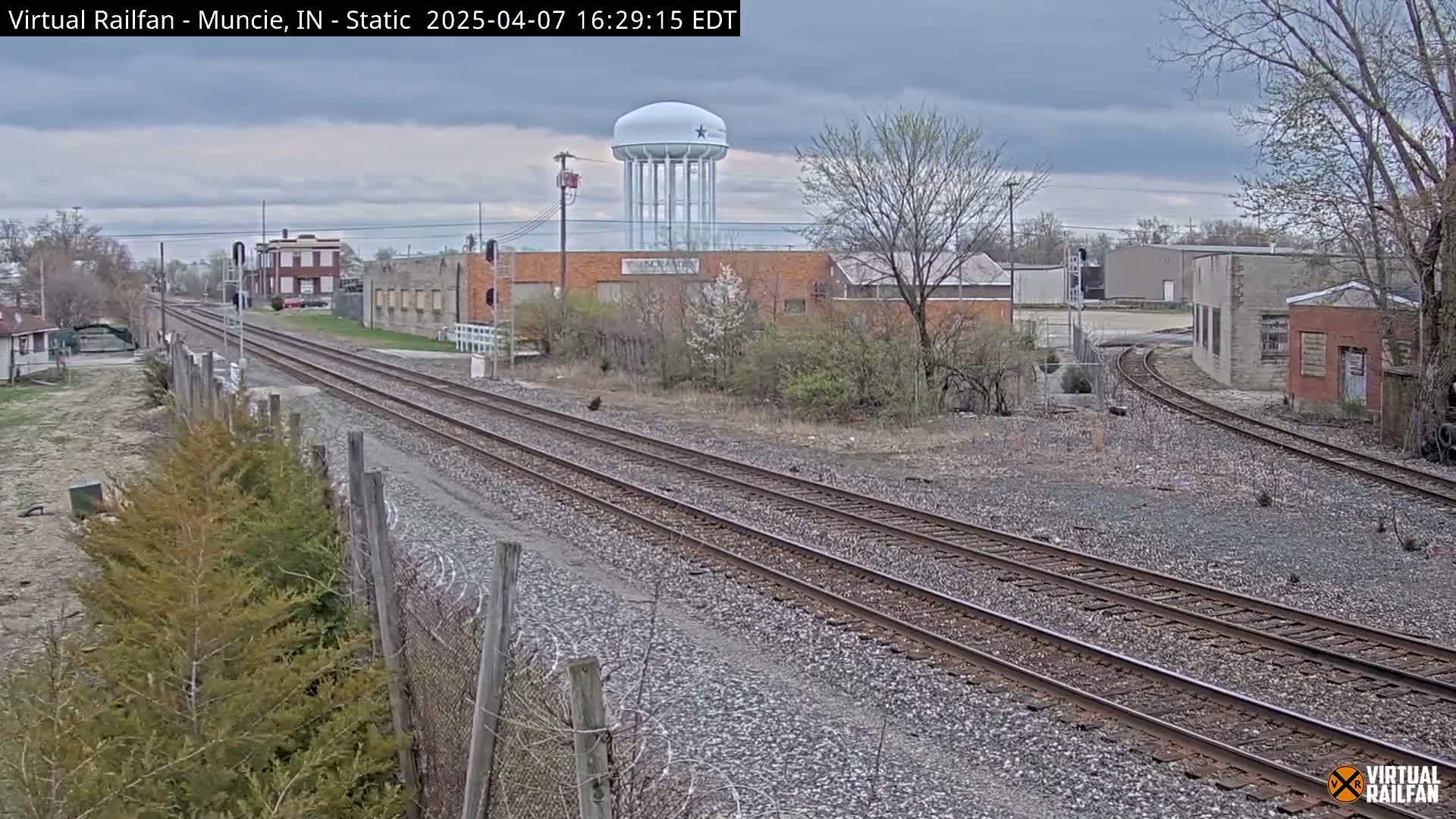 A view of railroad tracks extending into the distance, passing by industrial buildings and a water tower under an overcast sky.