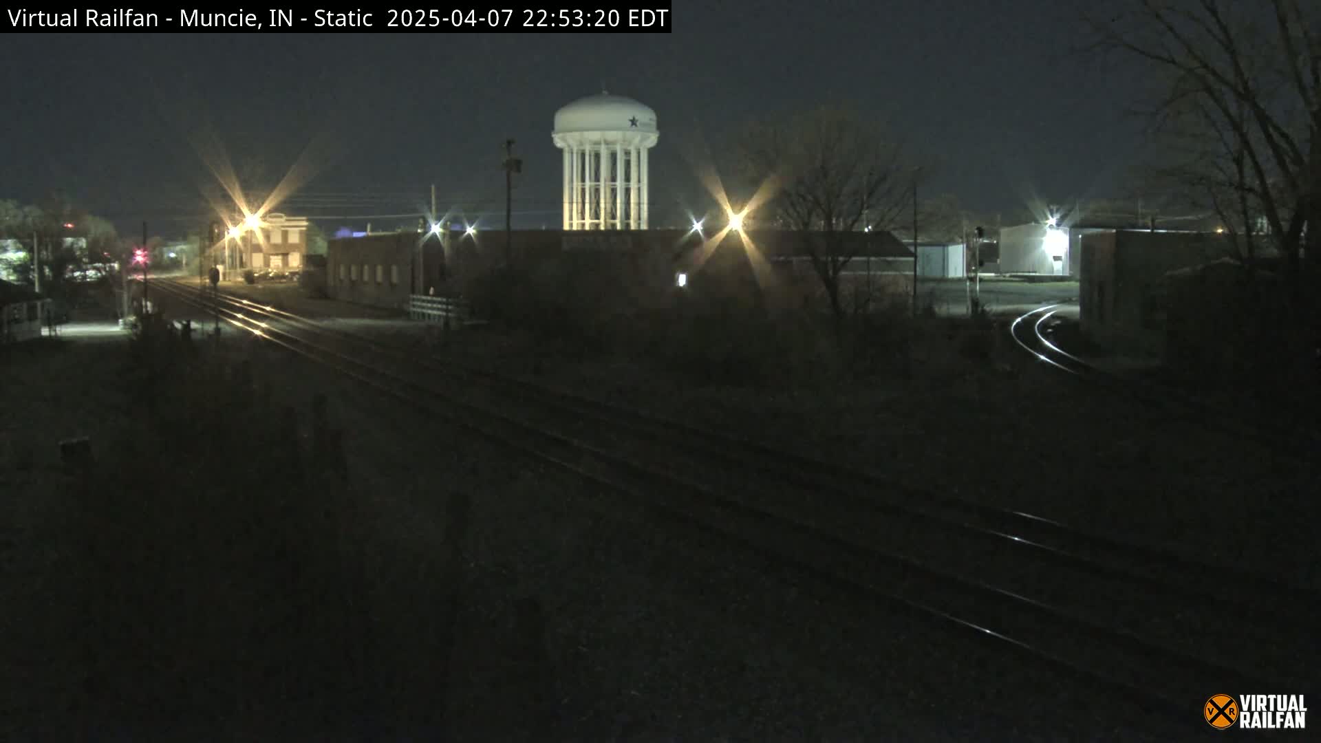 A nighttime view of railroad tracks leading toward a water tower and industrial buildings under a clear, dark sky.