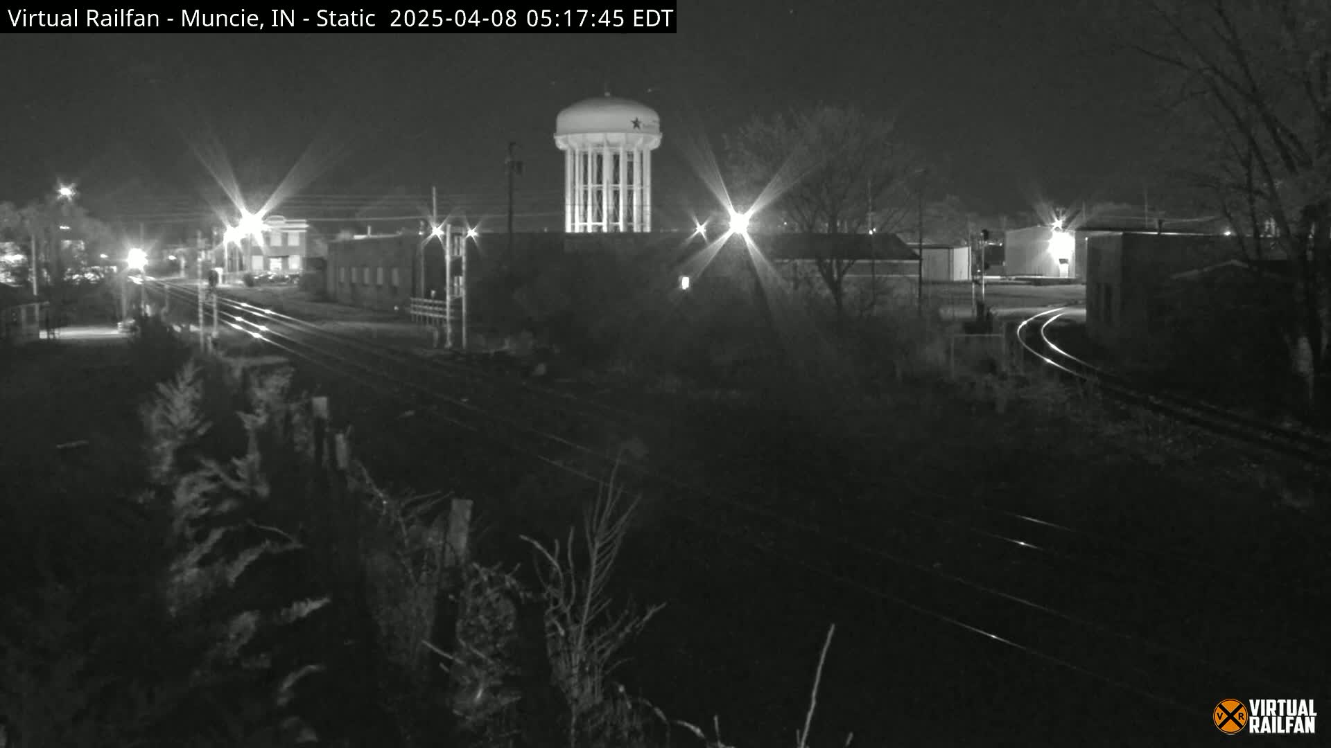 A nighttime, black-and-white view of railroad tracks curving past industrial buildings and a water tower under a dark sky.
