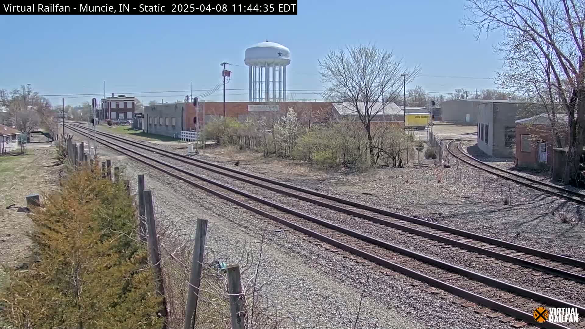Three sets of train tracks extend into the distance under a clear, sunny sky, past industrial buildings and a water tower, surrounded by leafless trees and shrubs.