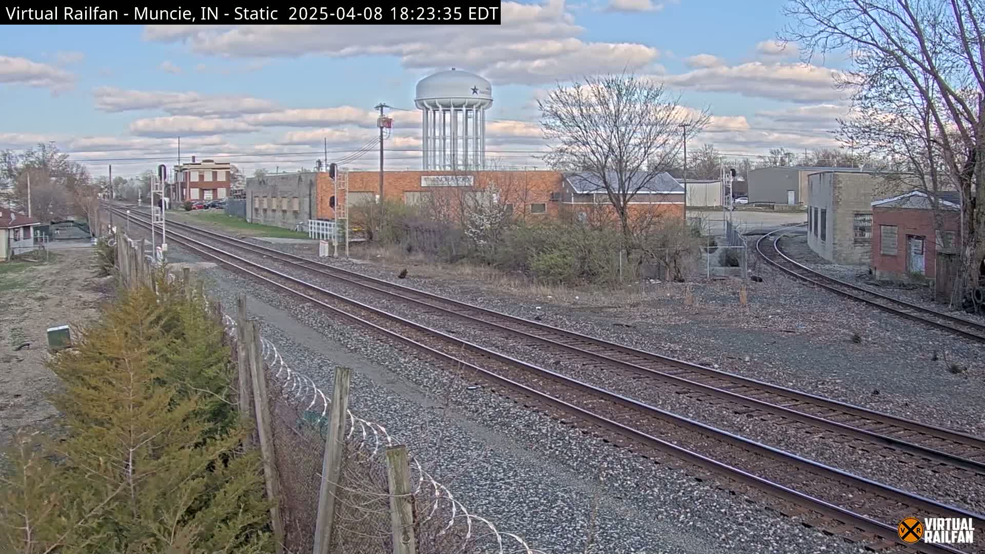 A view of railroad tracks splitting into two directions, passing industrial buildings and a water tower under a partly cloudy sky.
