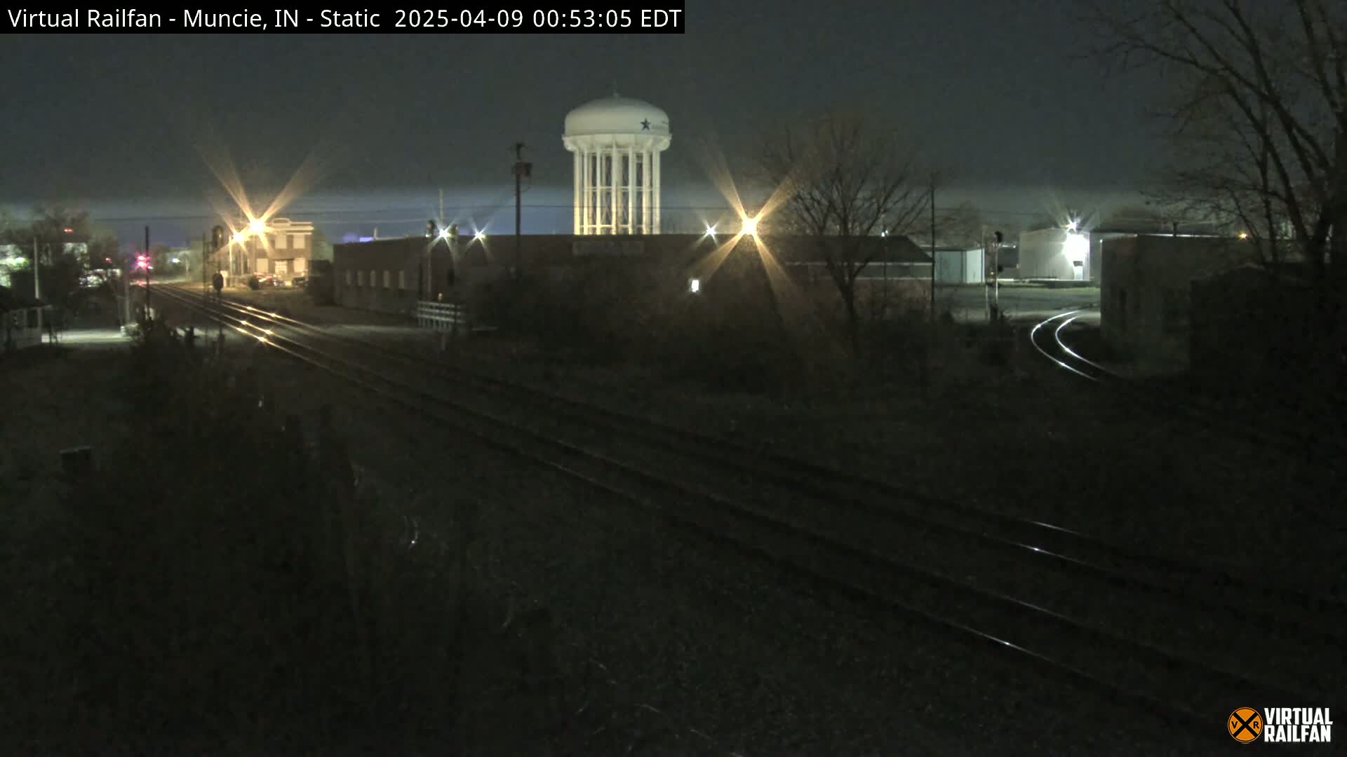 A nighttime view of railroad tracks, buildings, and a water tower under a dark sky.