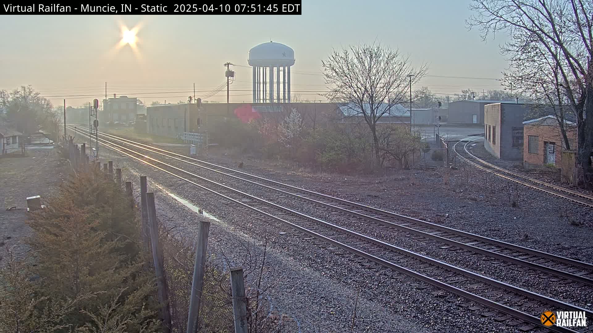 A hazy sunrise illuminates a view of multiple train tracks curving around industrial buildings and a water tower.