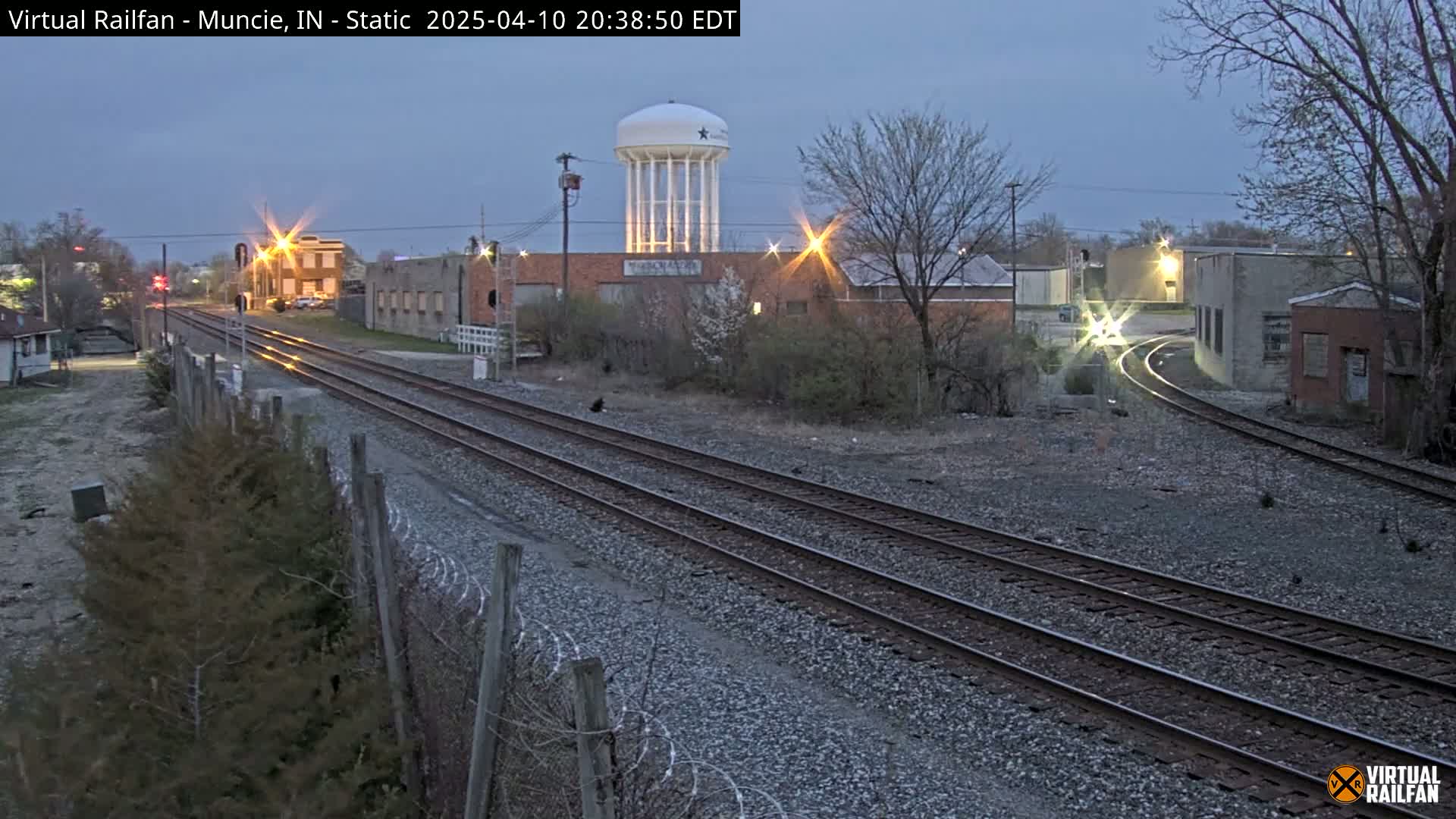 A twilight view of a railway track splitting into two directions, passing by industrial buildings and a water tower under a dusky sky.