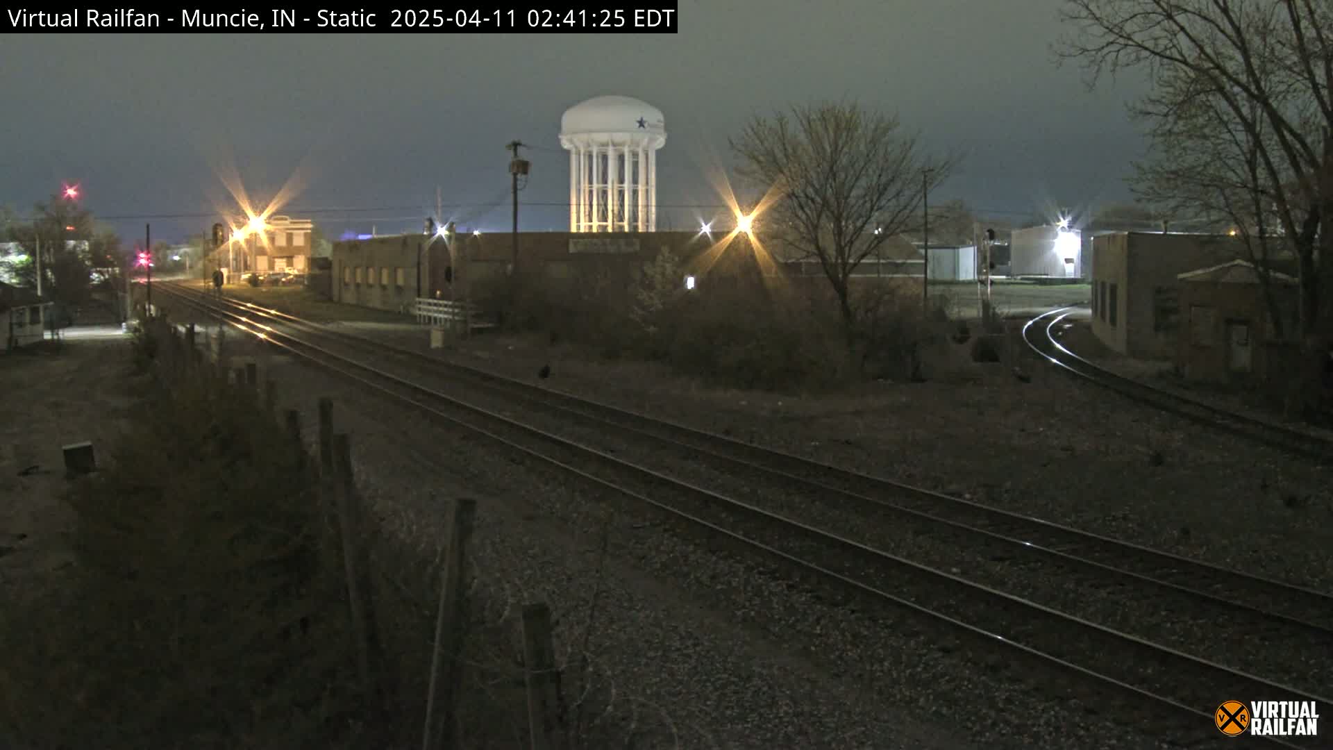 A nighttime view of railroad tracks, industrial buildings, and a water tower under a dark, overcast sky.