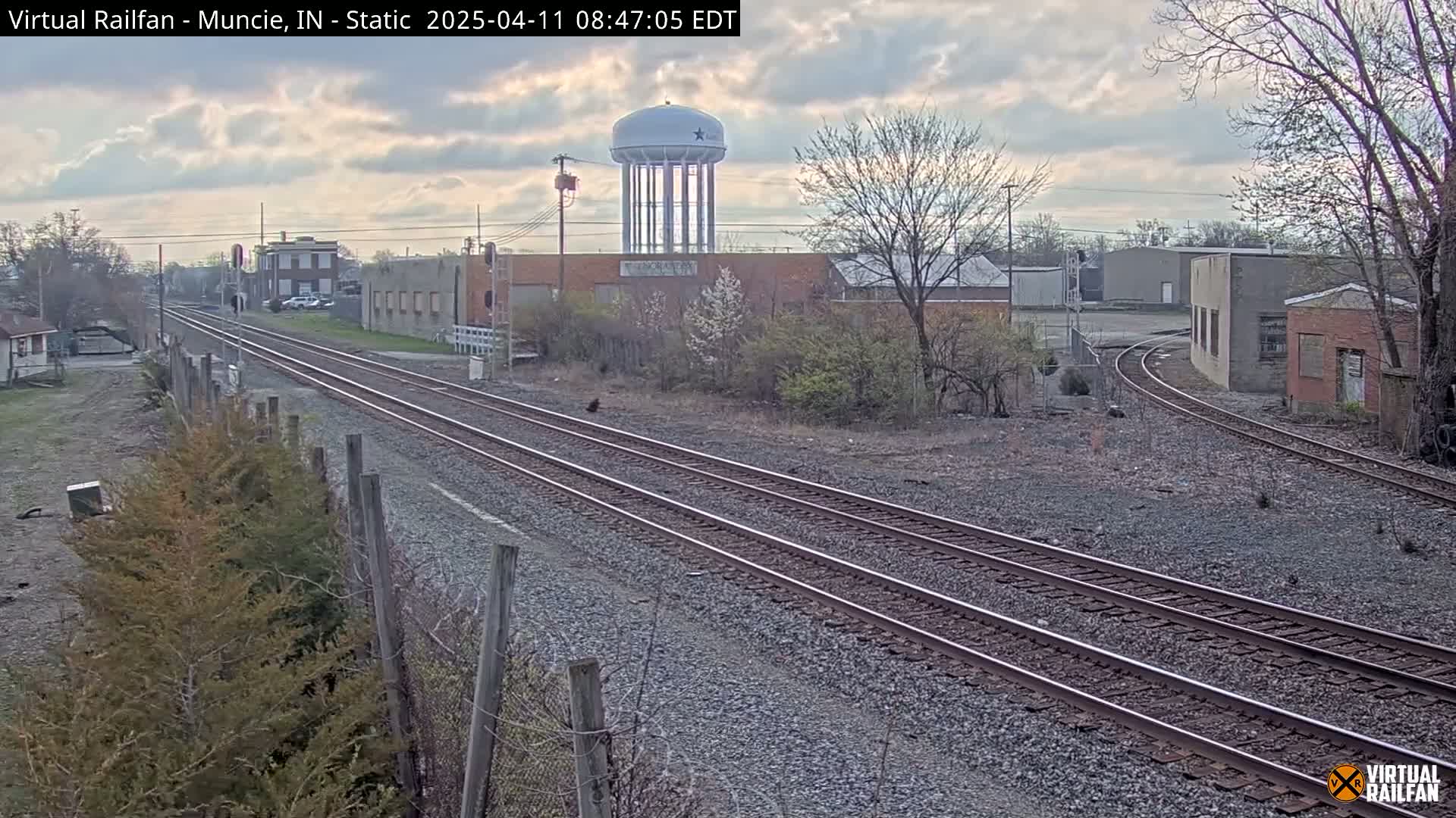 A view of railroad tracks extending into the distance under a partly cloudy sky, passing near industrial buildings and a water tower.