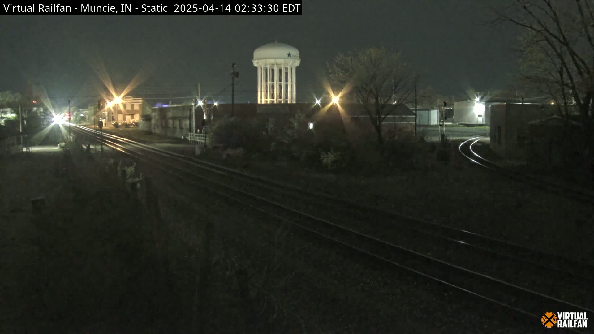A nighttime view of railroad tracks leading toward a water tower and buildings under a dark sky.