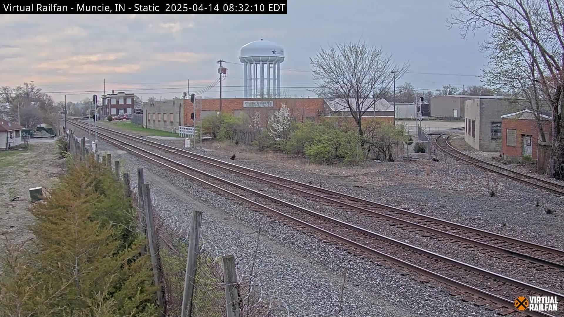 A view of railroad tracks splitting into two directions, passing by industrial buildings and a water tower under an overcast sky.