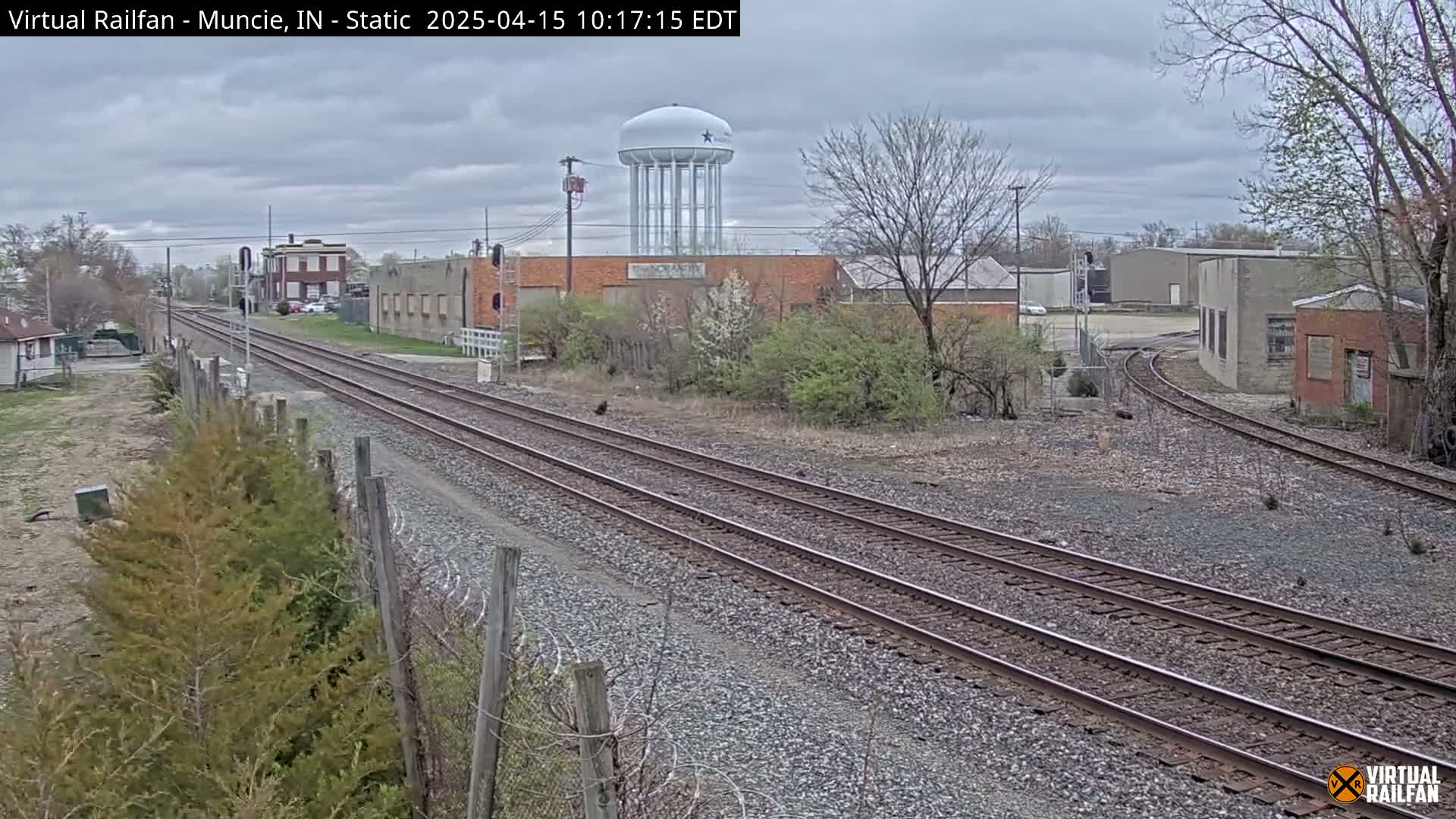 Two sets of train tracks extend into the distance past industrial buildings and a water tower under an overcast sky.