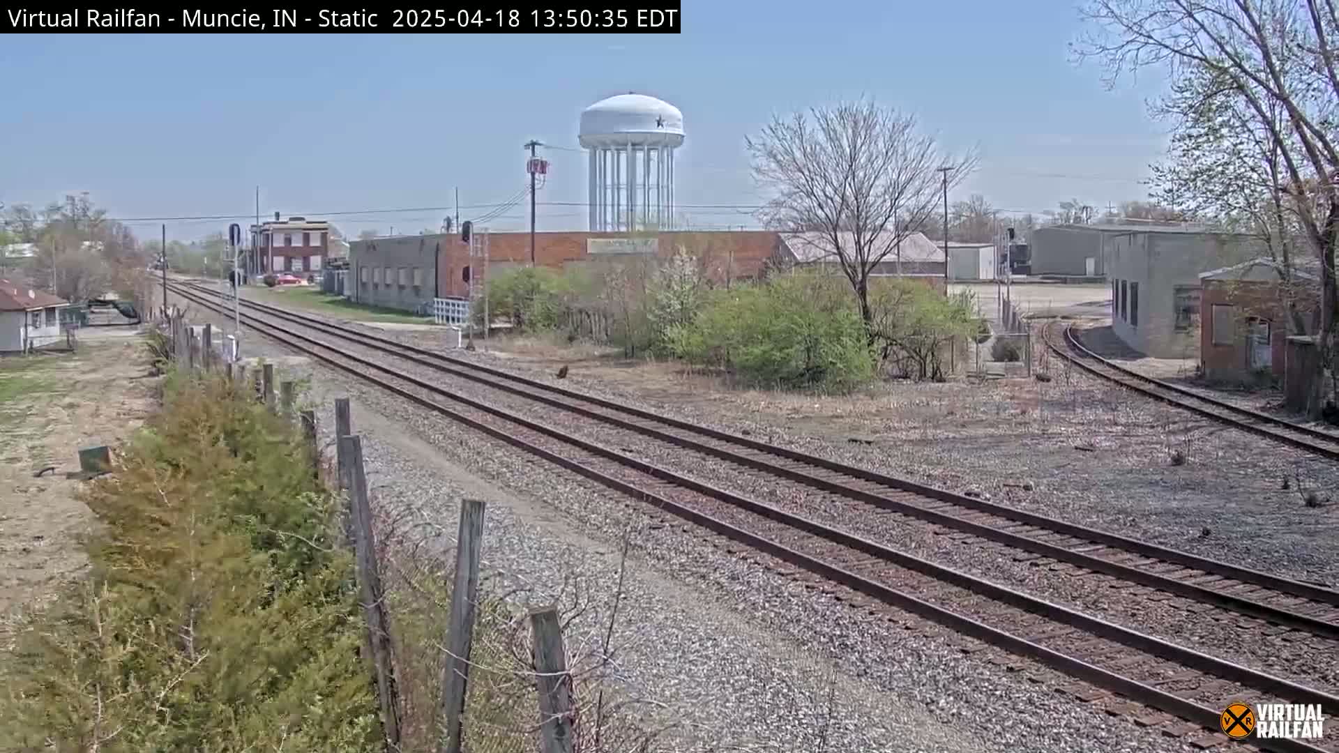A sunny day reveals railroad tracks extending into a distance, passing by industrial buildings, a water tower, and sparse vegetation.