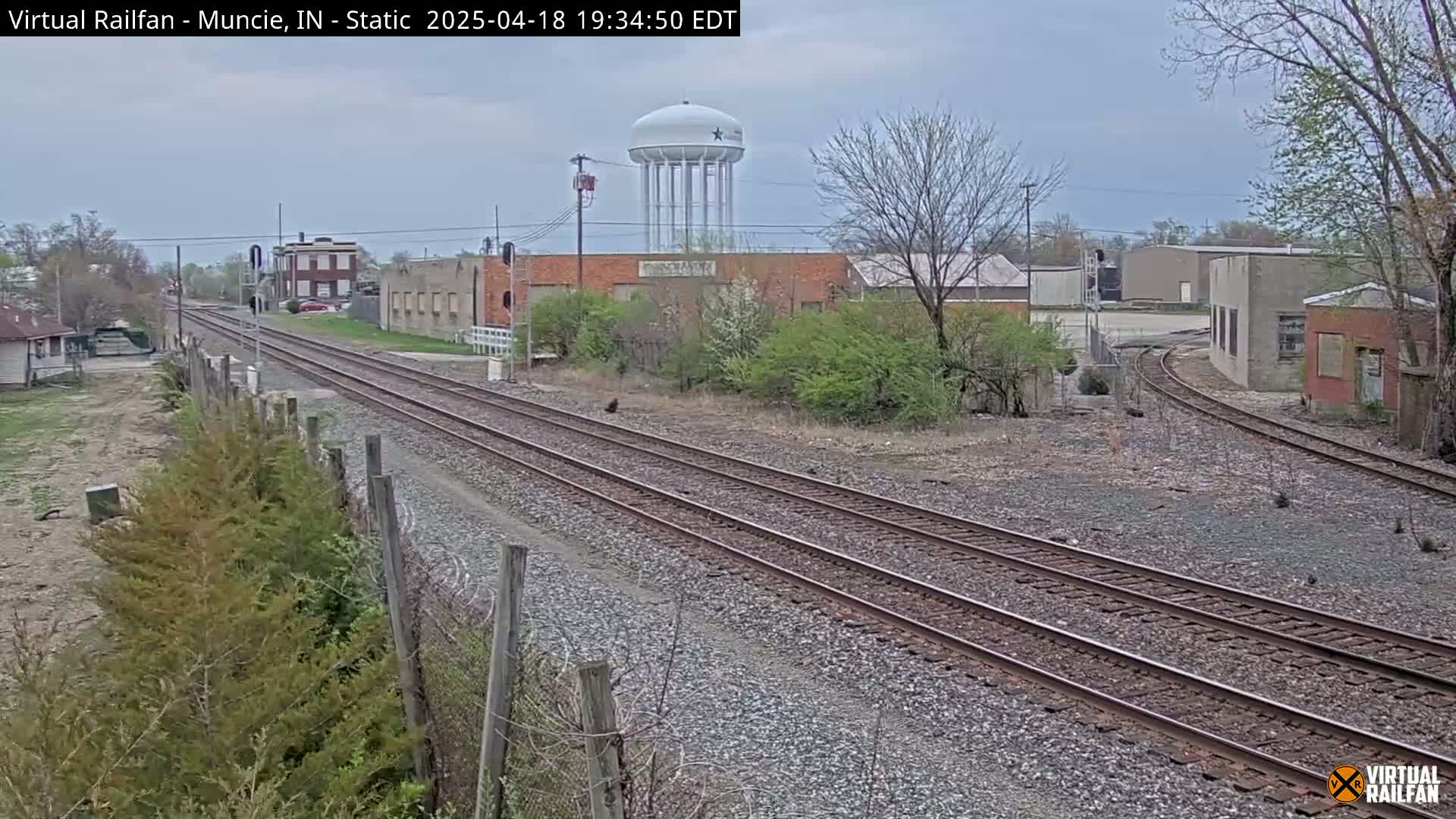 Two sets of train tracks curve through an industrial area under an overcast sky, past buildings and a water tower.