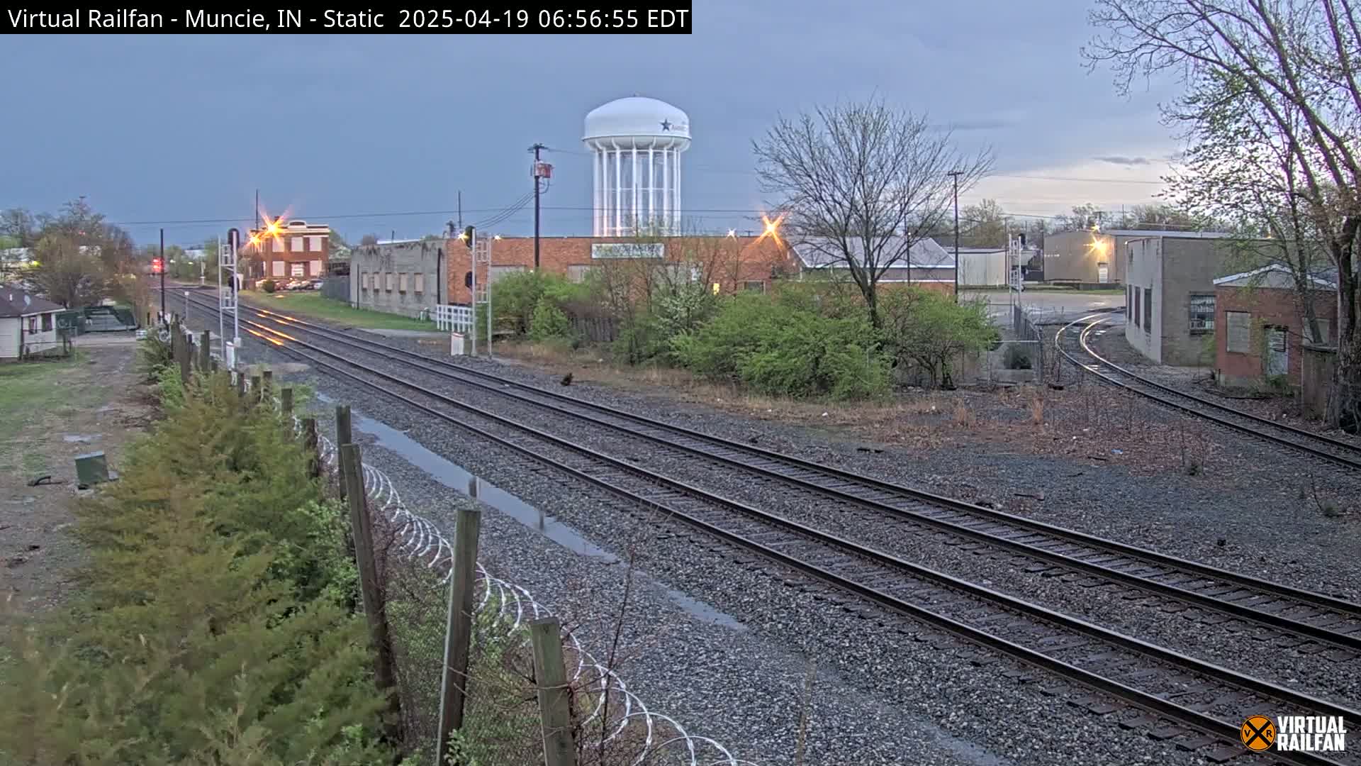 A view of railroad tracks extending into the distance past industrial buildings and a water tower under an overcast sky.