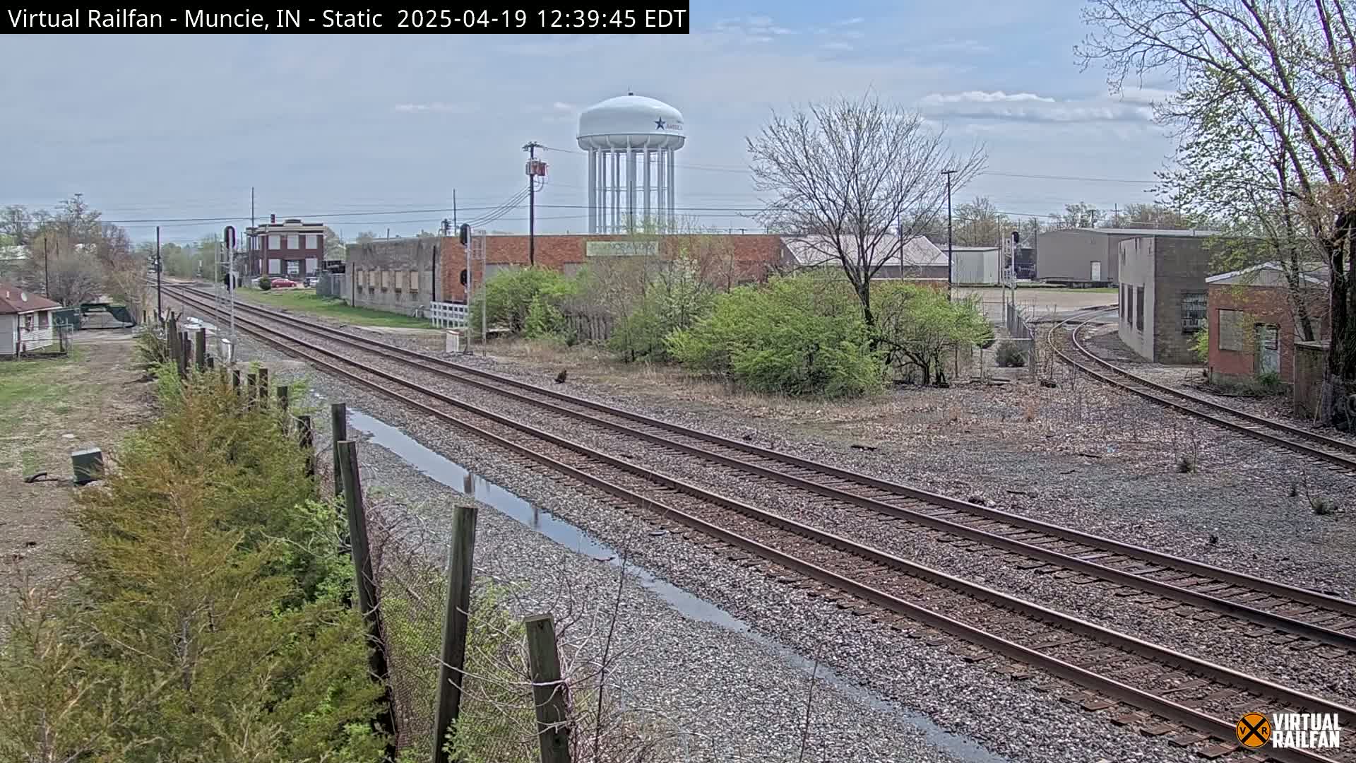 Two sets of train tracks stretch into the distance under a partly cloudy sky, passing alongside industrial buildings and a water tower.
