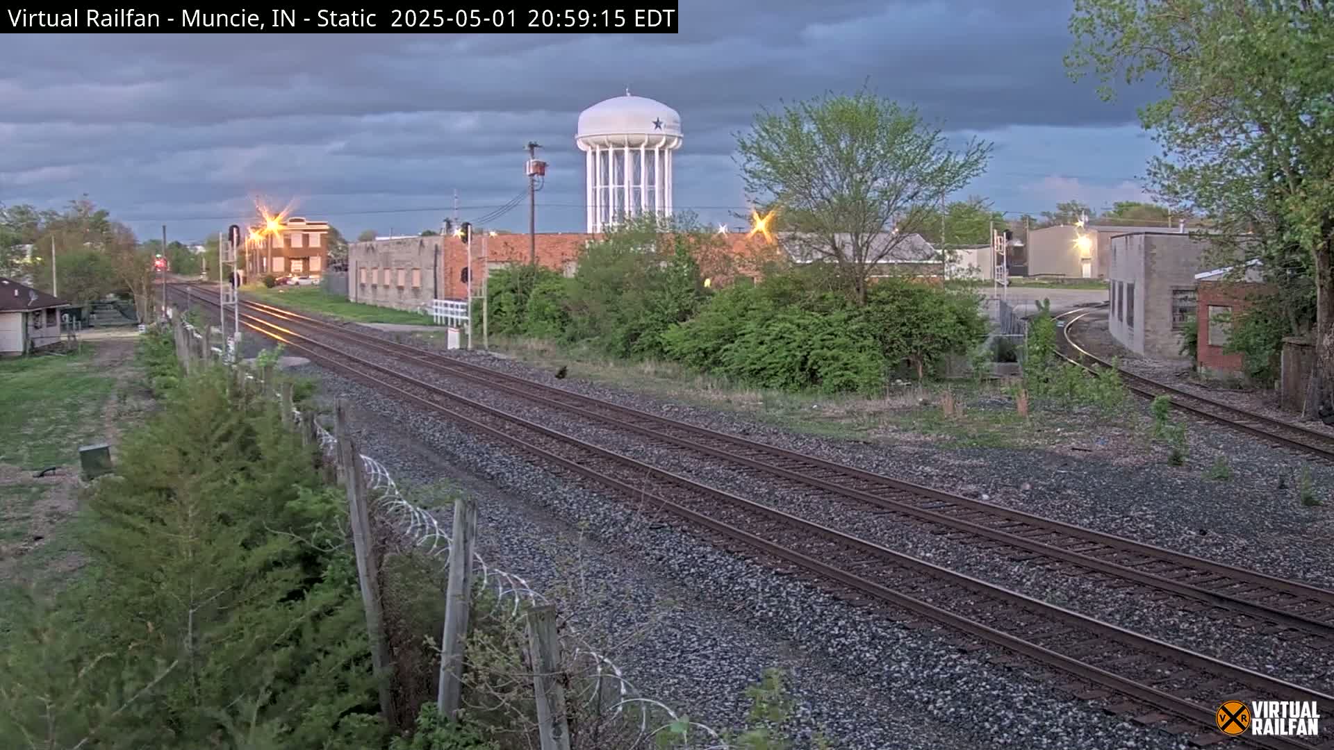 A view of railroad tracks under a partly cloudy evening sky, with industrial buildings and a water tower in the background.