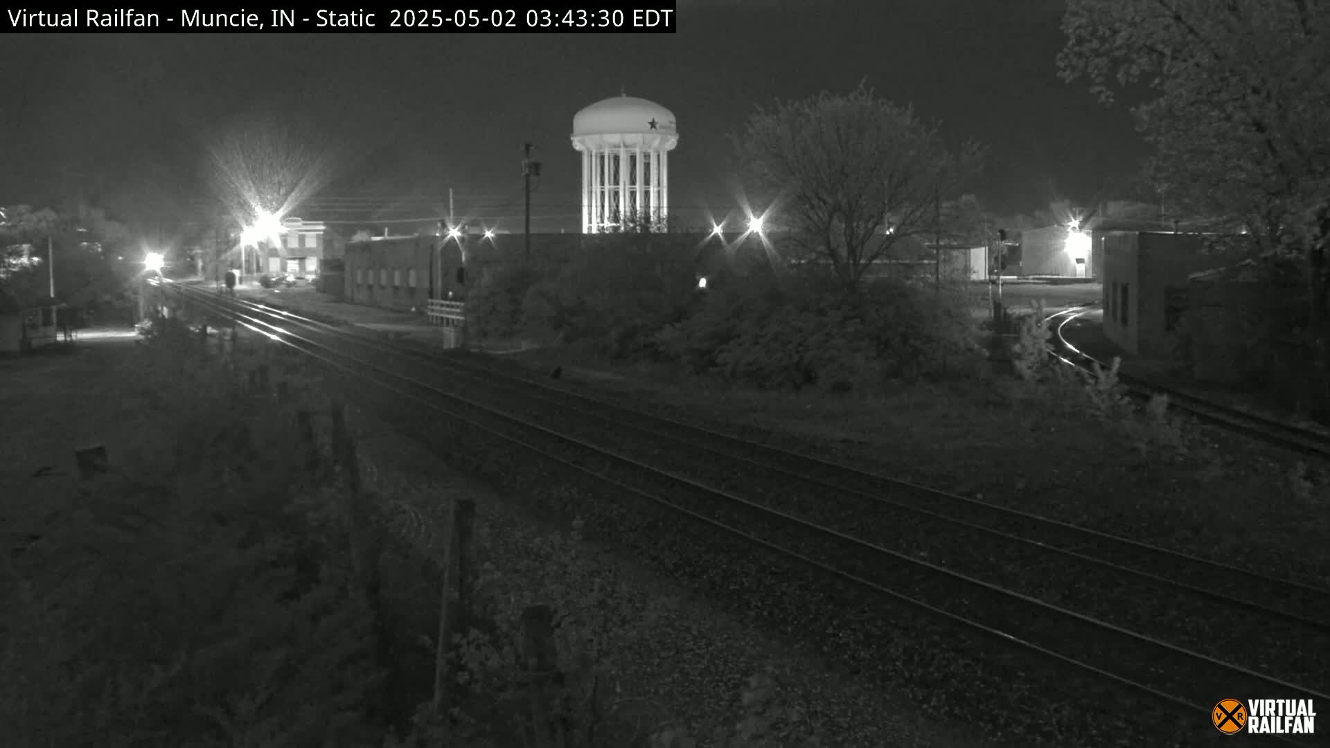 A nighttime, black-and-white view of railroad tracks extending toward a water tower and some industrial buildings under a dark sky.