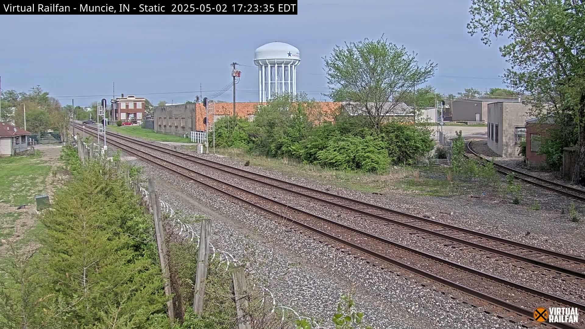 A view of railroad tracks under a mostly clear sky, with industrial buildings and a water tower visible in the background, surrounded by greenery.