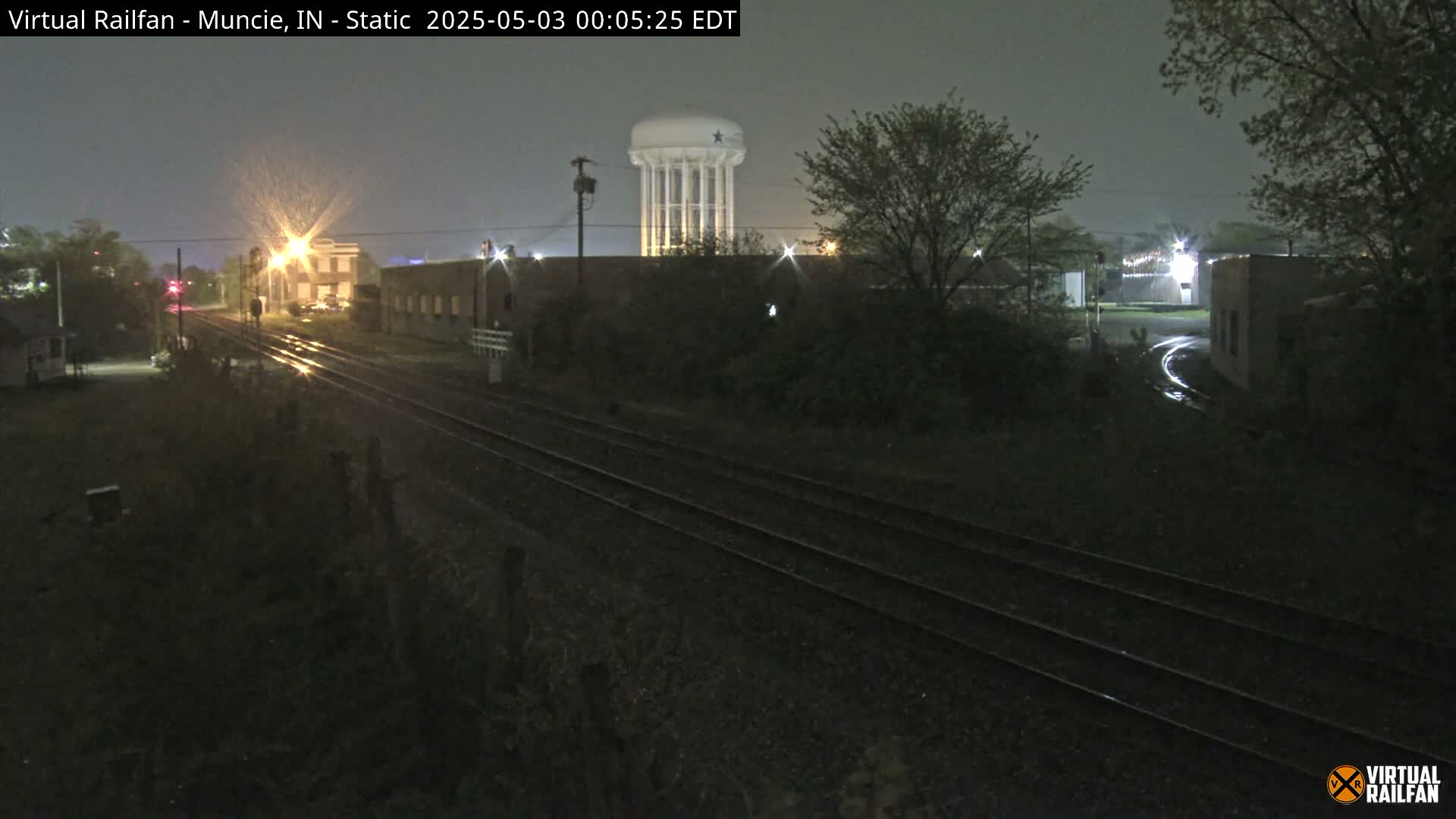 A nighttime view of rain falling on railroad tracks near a water tower and some buildings.