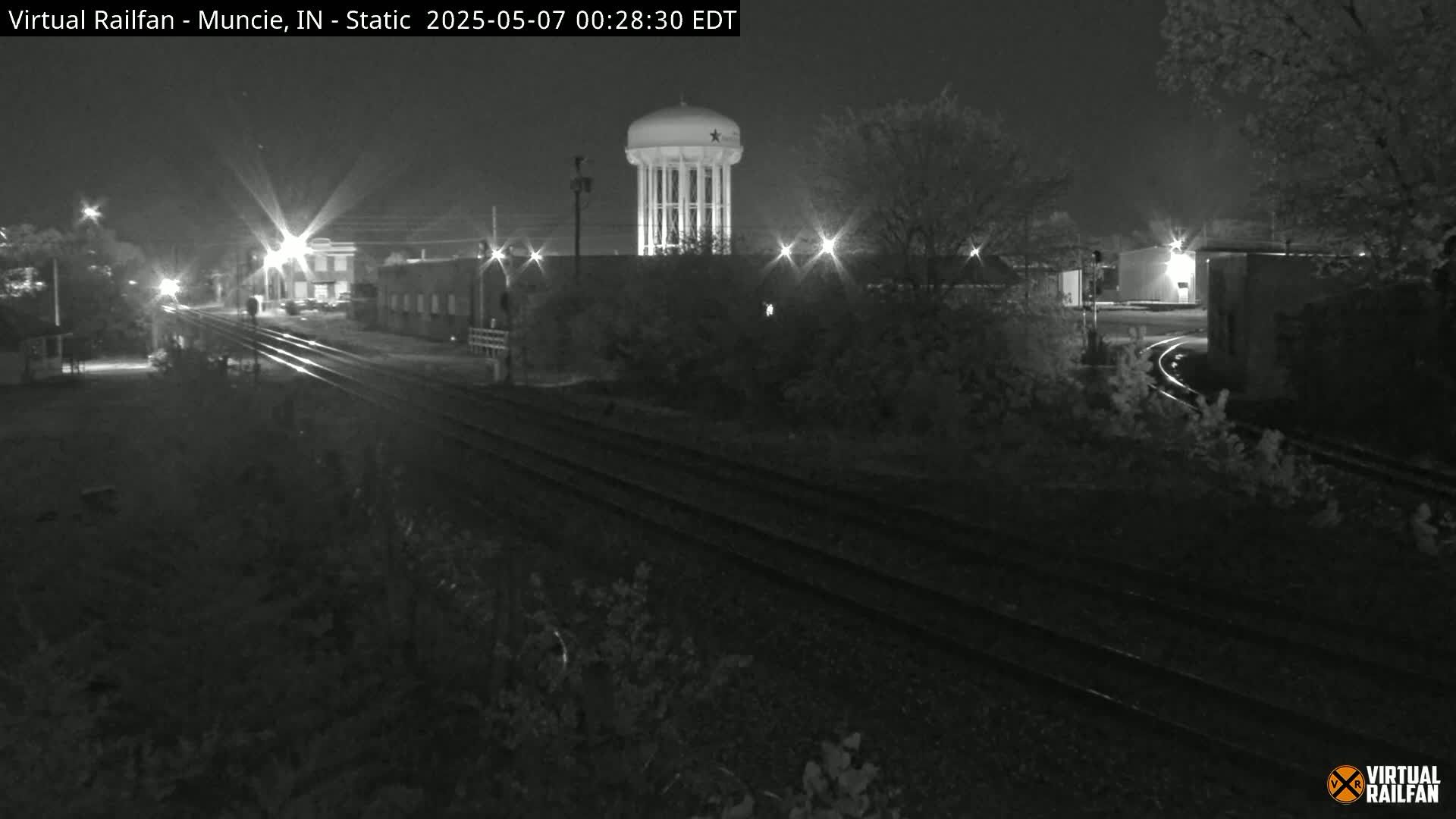 A nighttime, black-and-white view of railroad tracks leading toward a water tower, with streetlights illuminating the surrounding area.