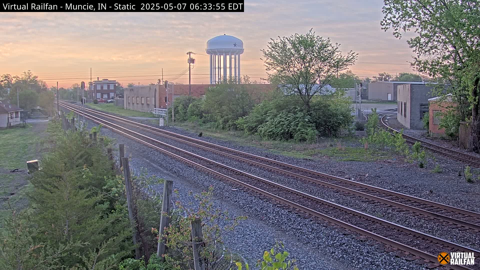 Under a soft, dusky sky, three sets of train tracks extend into the distance, passing industrial buildings and a water tower, surrounded by budding greenery.