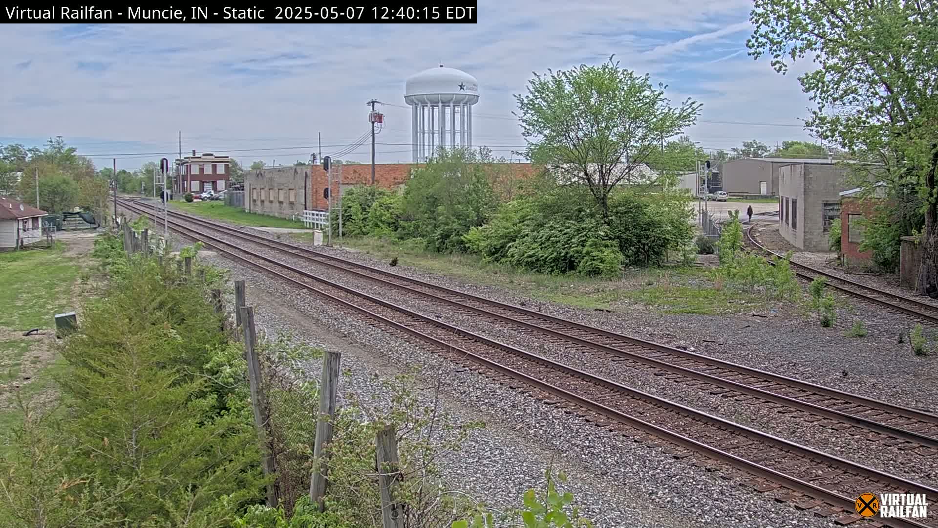Three sets of train tracks run through a town on a partly cloudy day, past industrial buildings and a water tower.
