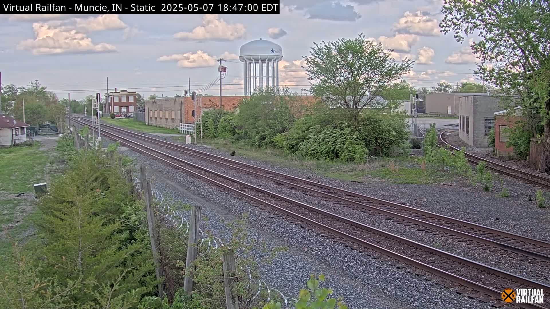 Railroad tracks extend into the distance past industrial buildings and a water tower under a partly cloudy sky.