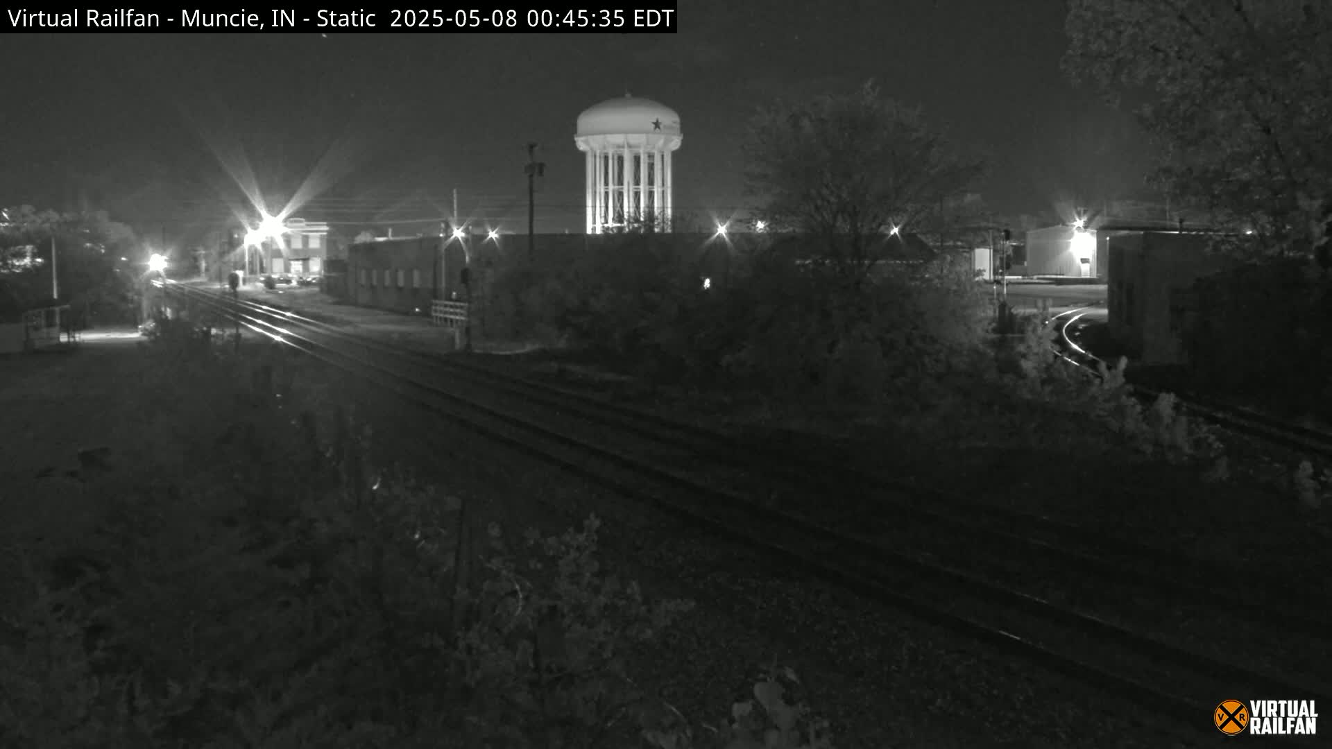 A nighttime view of railroad tracks leading toward a water tower, surrounded by dark buildings and vegetation.