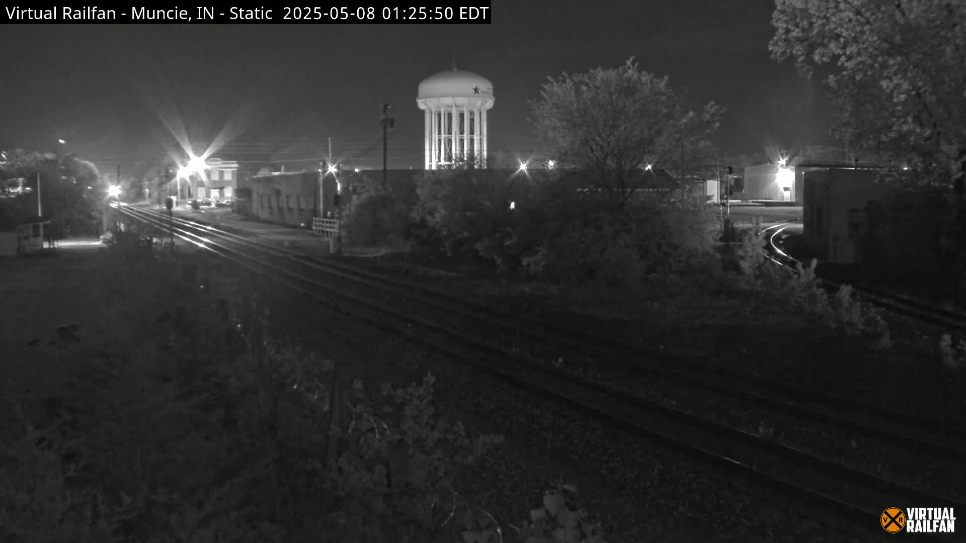 At night, railroad tracks extend from the foreground into the distance, passing under a water tower and alongside buildings, all under a dark sky.