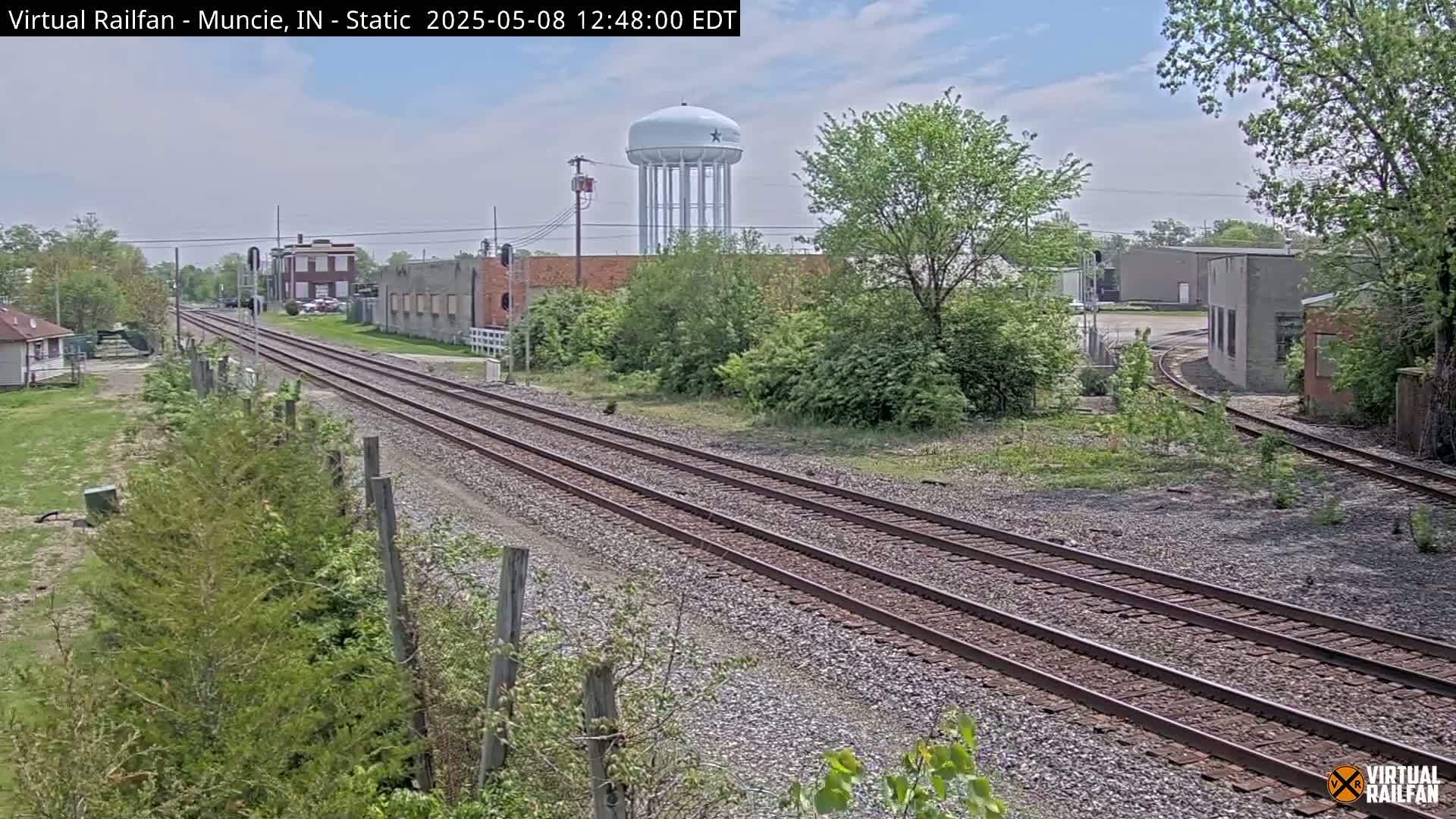 A view of railroad tracks on a partly sunny day, with industrial buildings and a water tower in the background, surrounded by green vegetation.