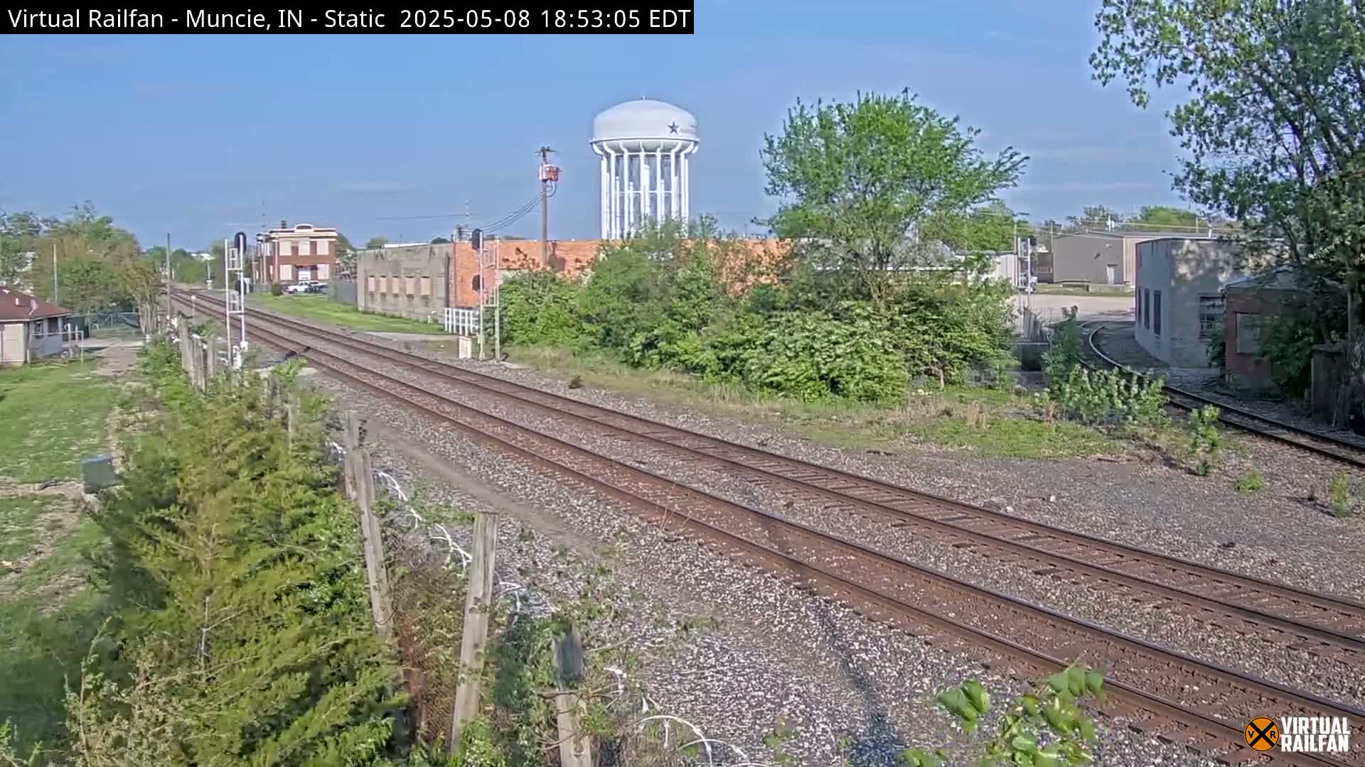 A view of a railway track extending into the distance under a clear sky, past industrial buildings and a water tower, with lush green vegetation alongside.