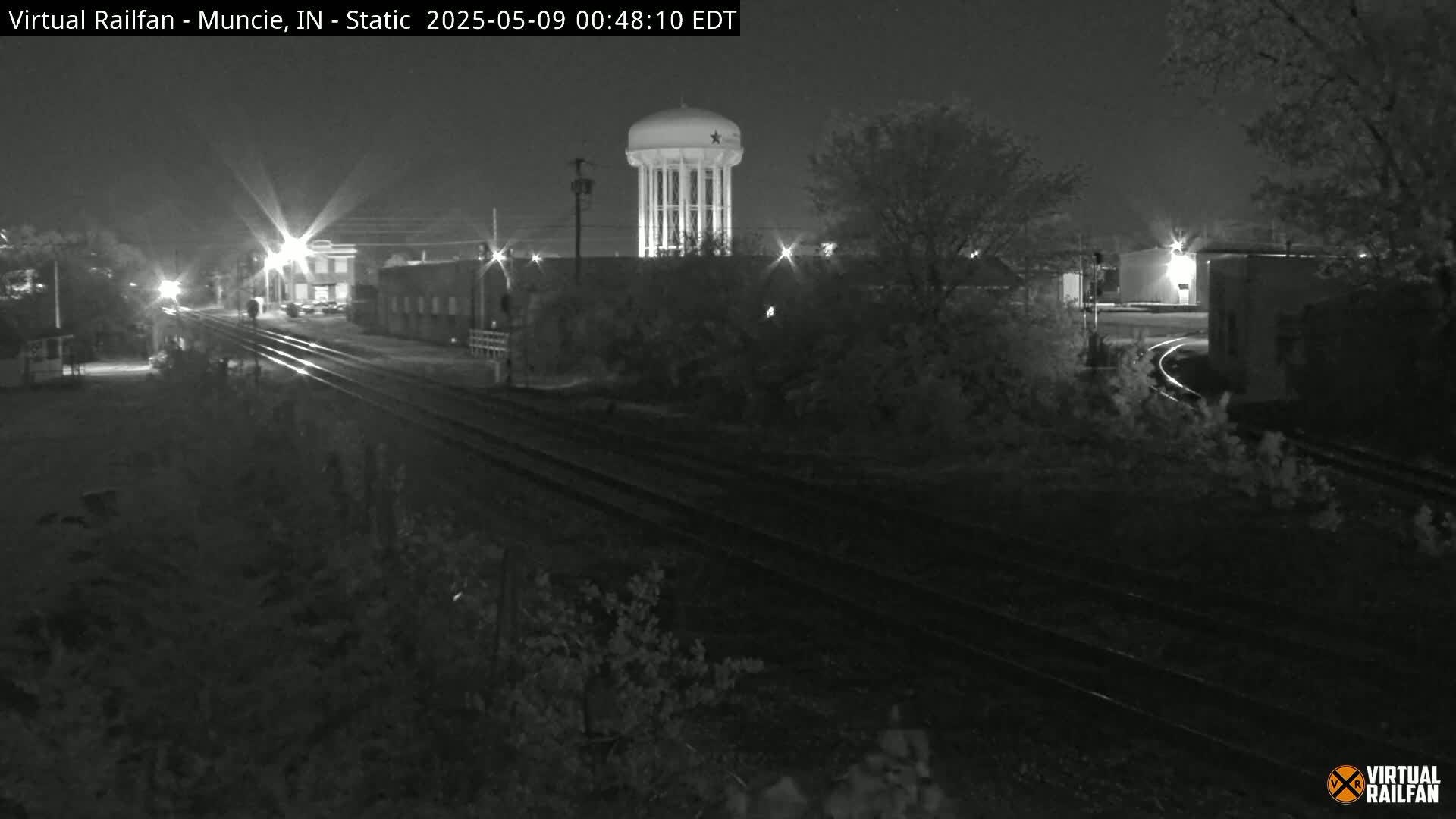 A nighttime, black-and-white view of railroad tracks extending toward a water tower and industrial buildings under a clear, dark sky.
