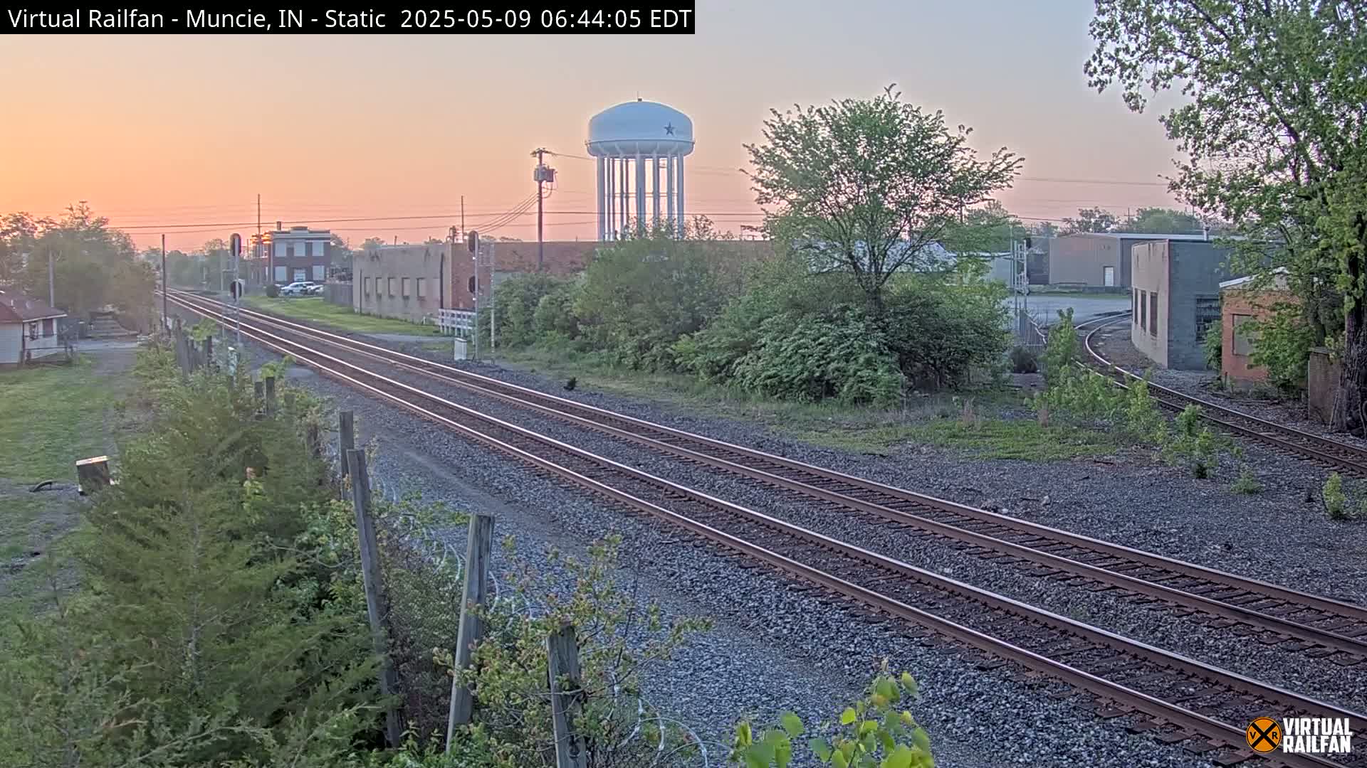 Triple train tracks extend into the distance at sunrise past industrial buildings and a water tower, under a pale pink sky.
