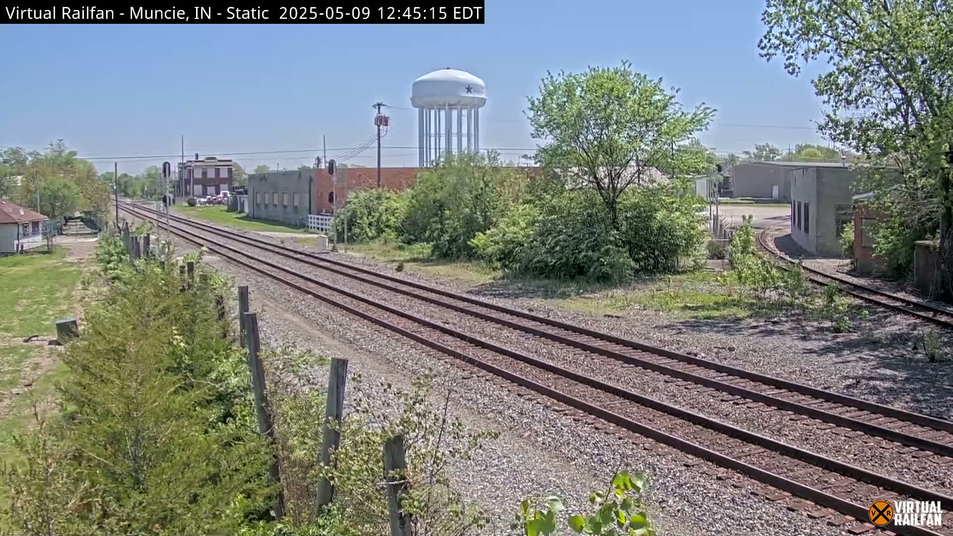 Under a bright, sunny sky, railroad tracks stretch into the distance past industrial buildings and a water tower, surrounded by greenery.