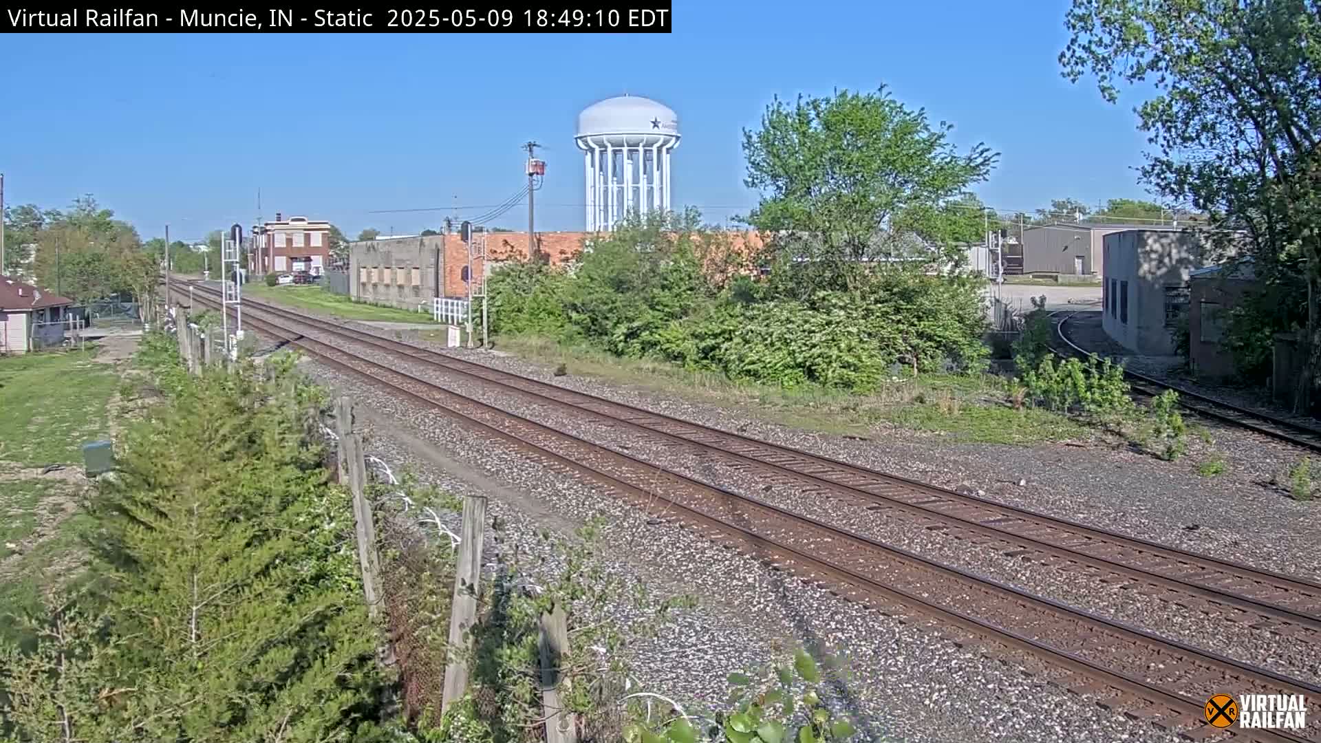 Under a clear blue sky, a view of railroad tracks stretches into the distance, passing by industrial buildings and a water tower.
