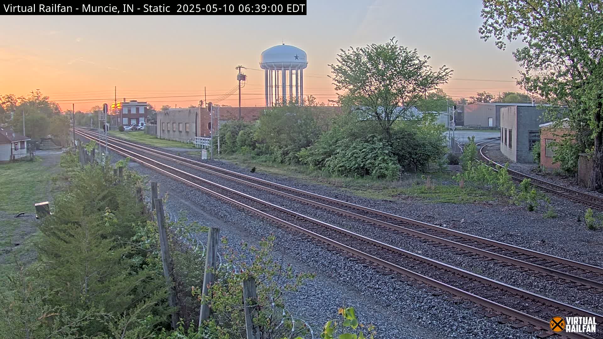 At sunrise, three sets of train tracks extend into the distance, past industrial buildings and a water tower, amidst budding green vegetation.