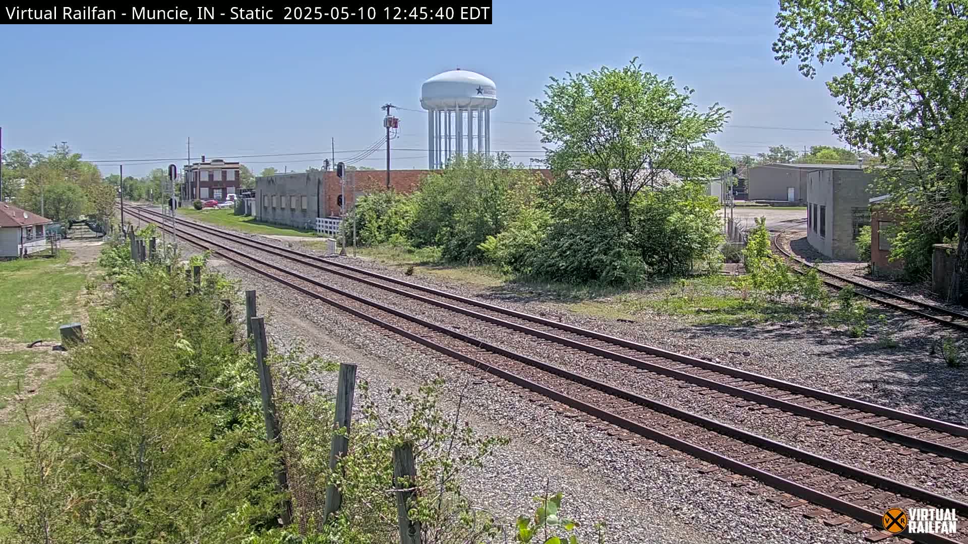 A sunny day view of train tracks splitting into two, with industrial buildings and a water tower visible in the background.